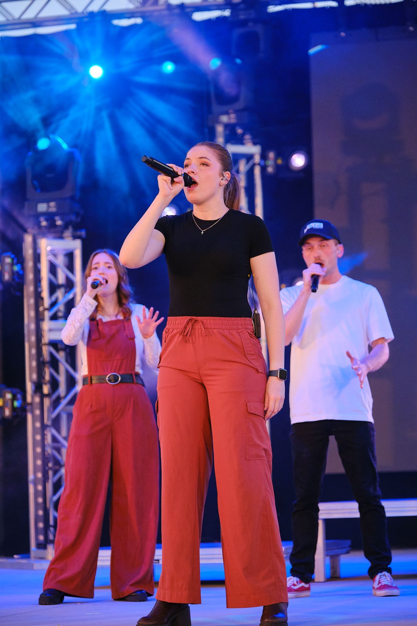 Three performers sing into microphones on a stage with bright blue spotlights and metal trusses in the background.