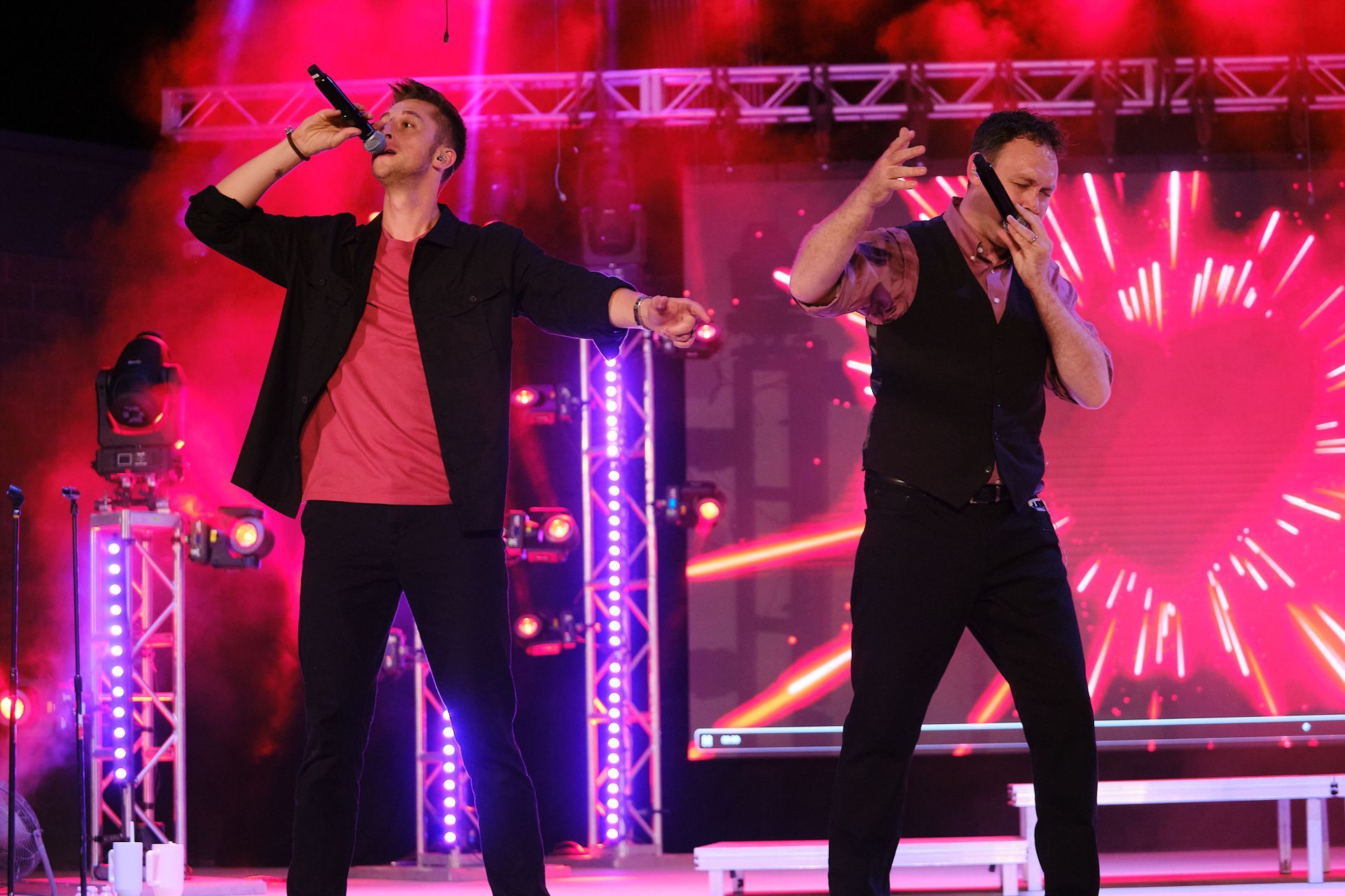Two performers on a stage under bright pink lights, singing into microphones during a live event.