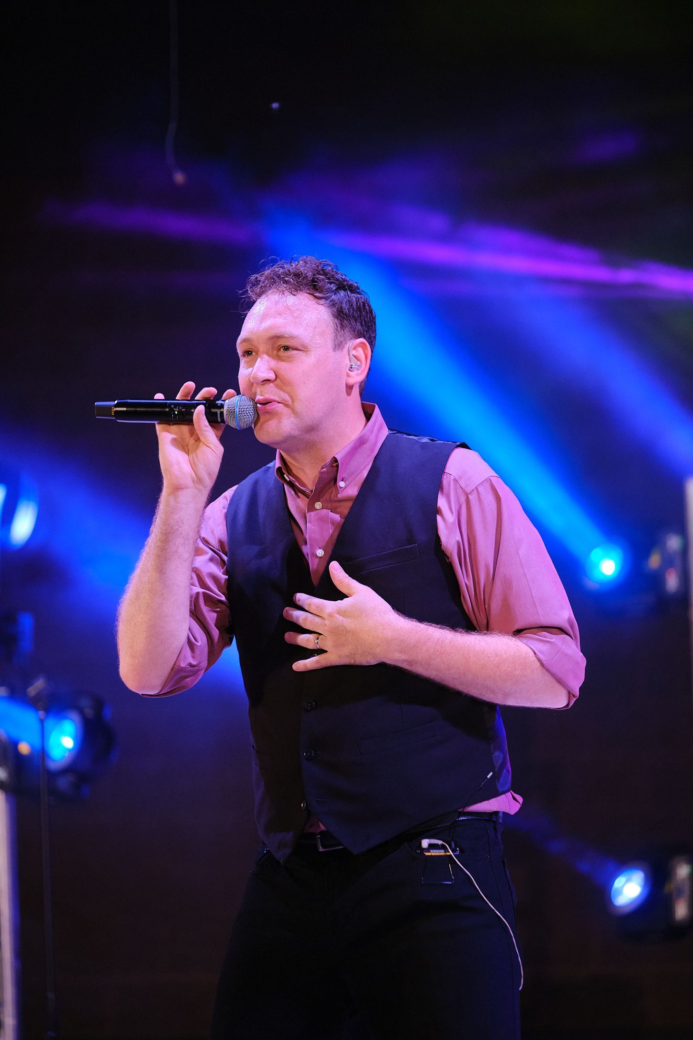 A person sings into a microphone on stage, wearing a mauve button-down shirt and black vest under bright blue spotlights.