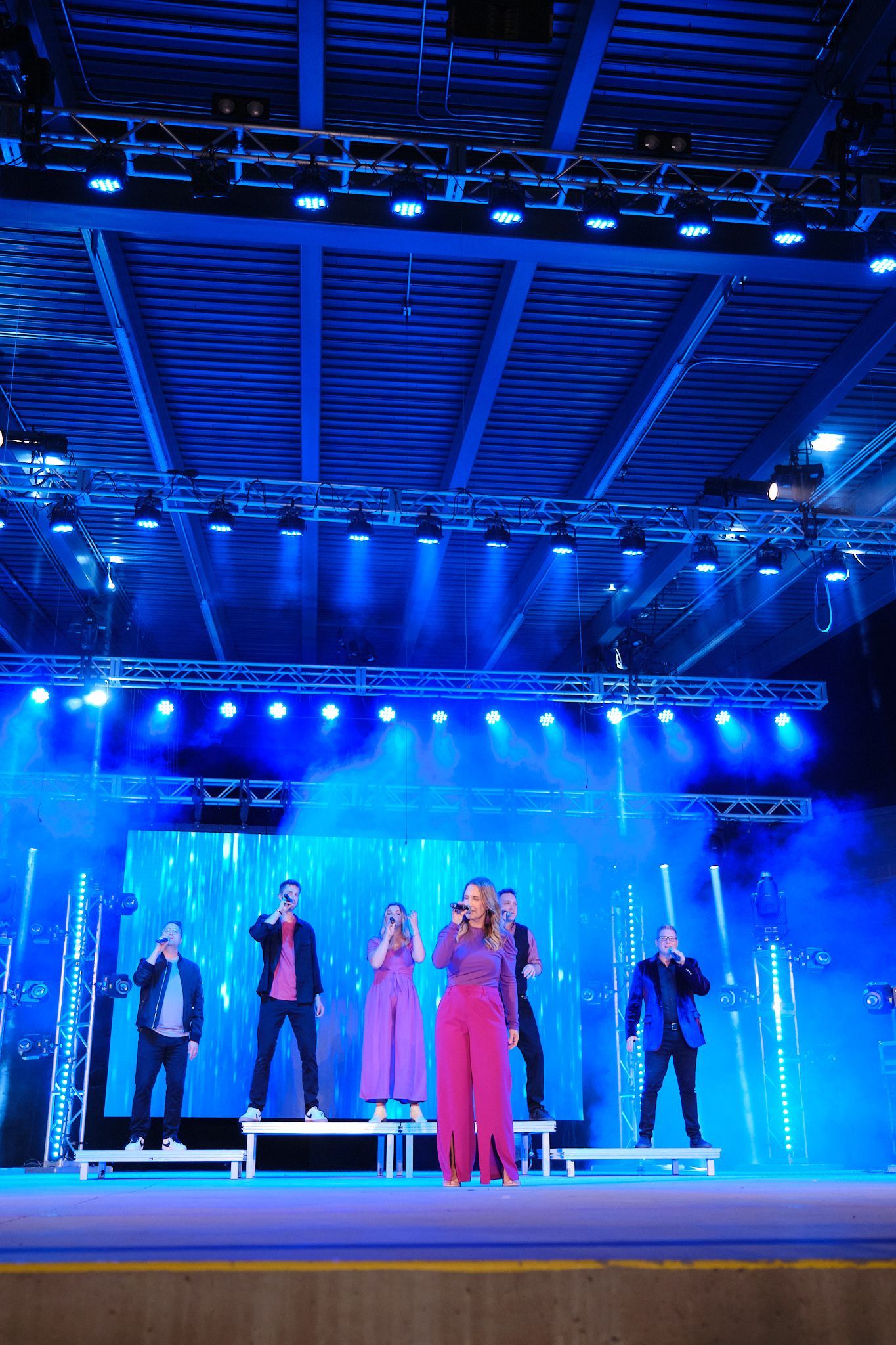A group of six people stand on a tiered stage, performing under bright blue spotlights in a large, dark venue.