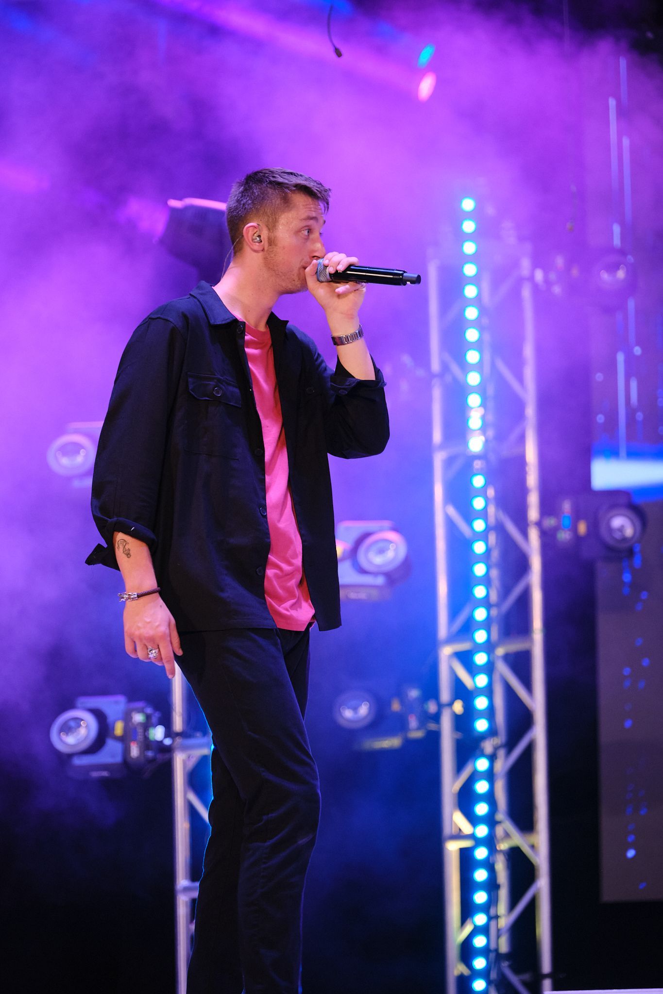 A performer in a black shirt and pink undershirt sings into a microphone on a stage with purple lighting and metal rigging.