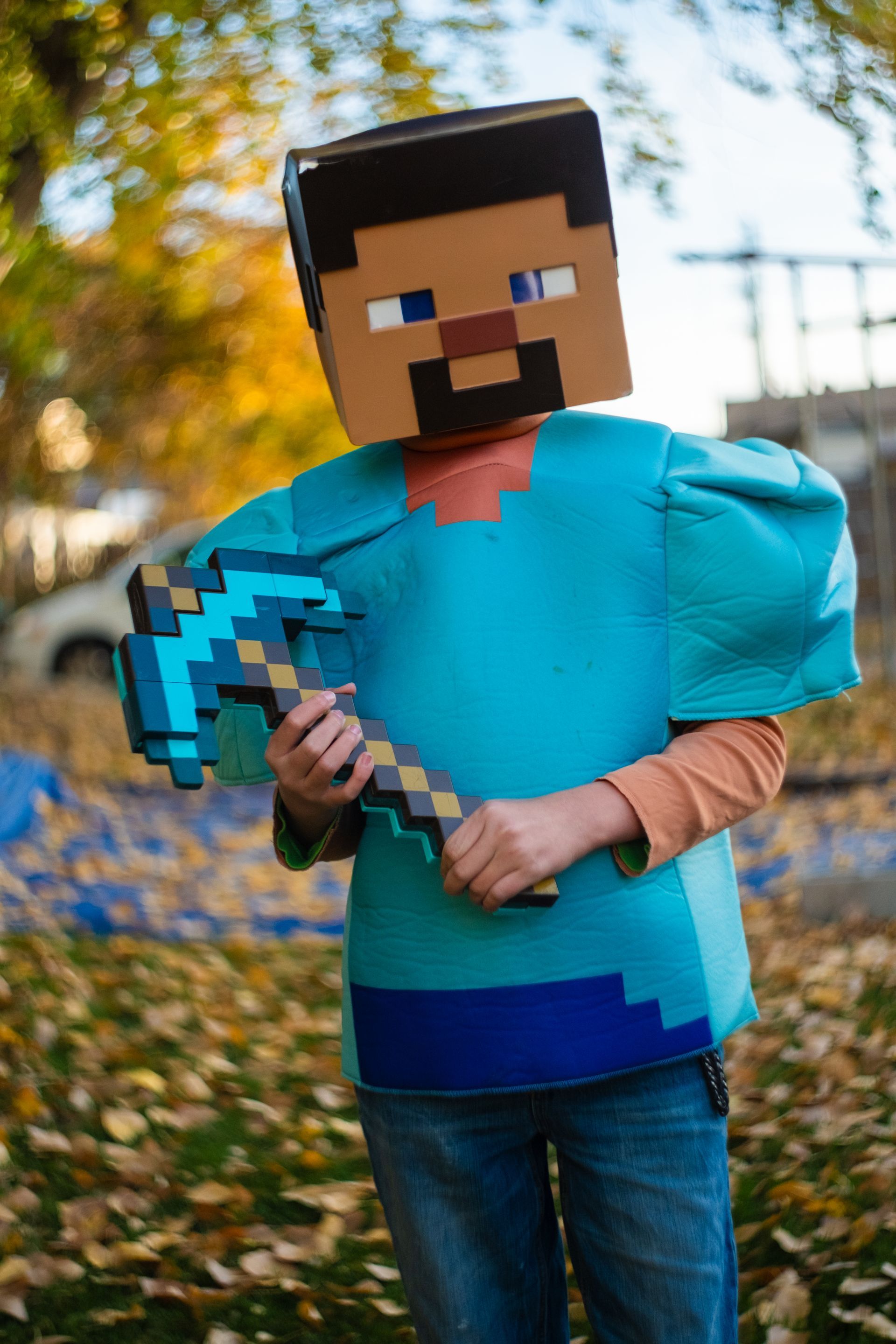 A child in a Minecraft Steve costume with a box head and a pixelated blue pickaxe, standing outdoors in fall leaves.