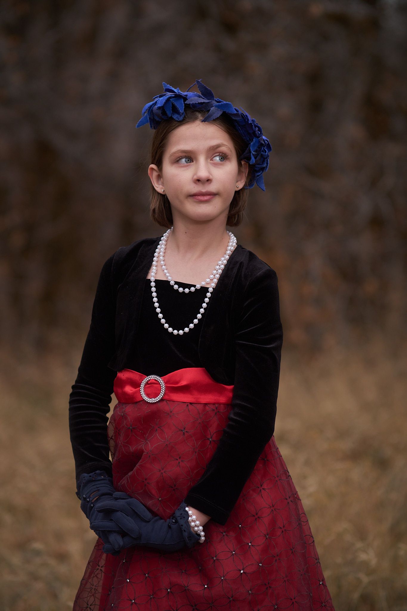A person wearing a dark velvet bolero, a red embellished skirt, a blue headpiece, and pearl necklaces stands outdoors.