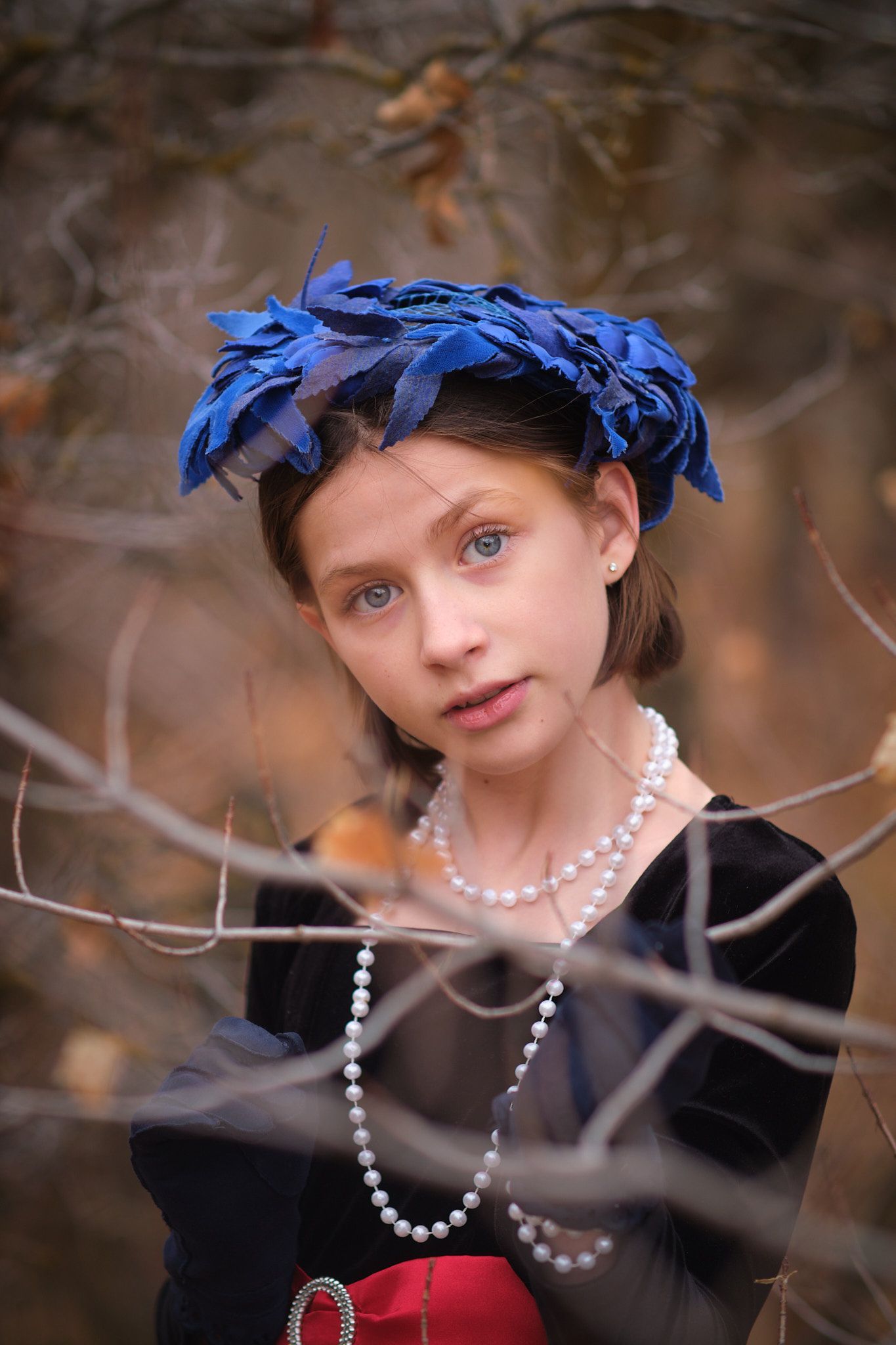 A person wearing a blue feathered hat, layered pearl necklaces, and a black top, framed by blurred brown tree branches.