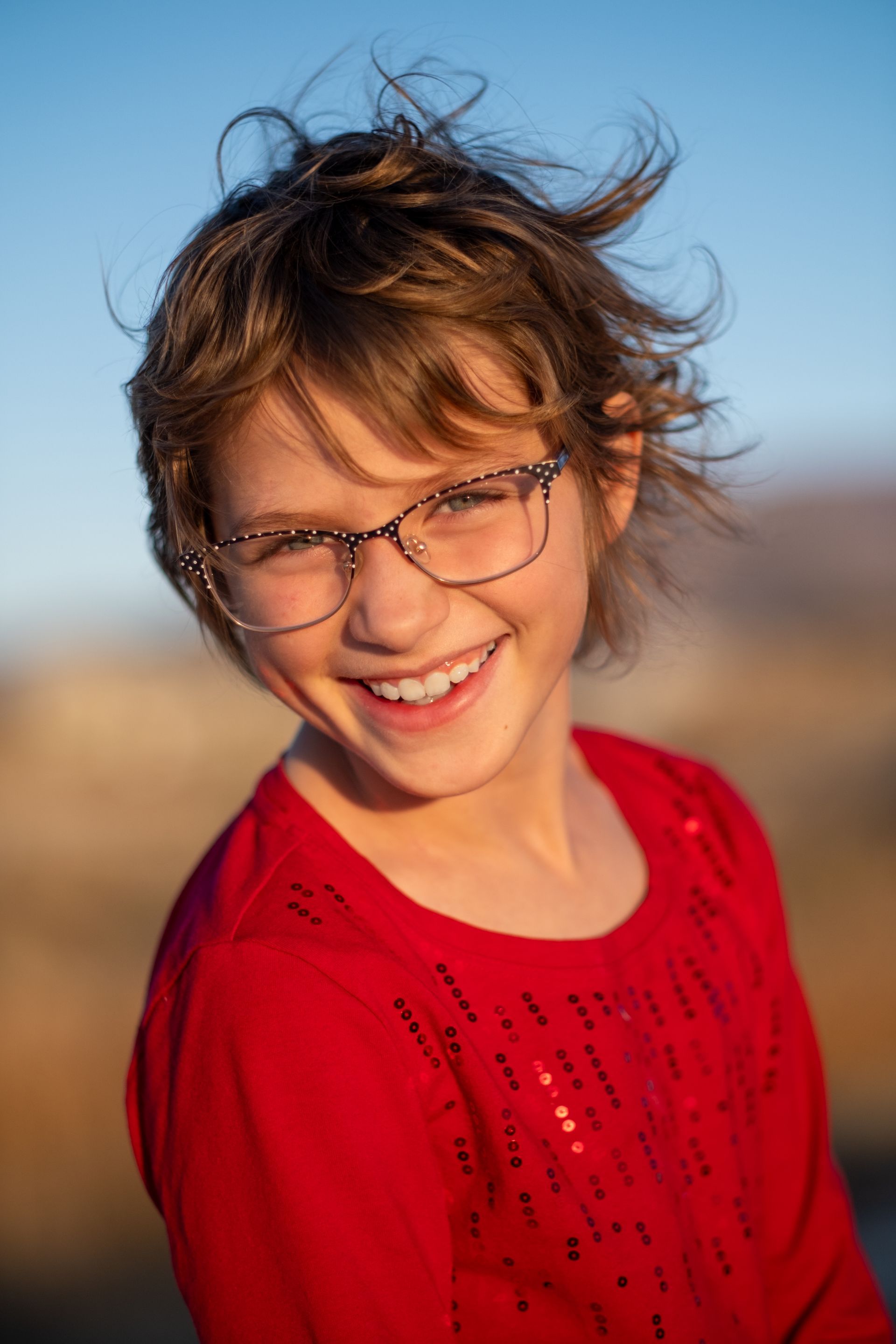 A smiling person with messy brown hair and glasses wears a red shirt outdoors against a blurred, sunny landscape.
