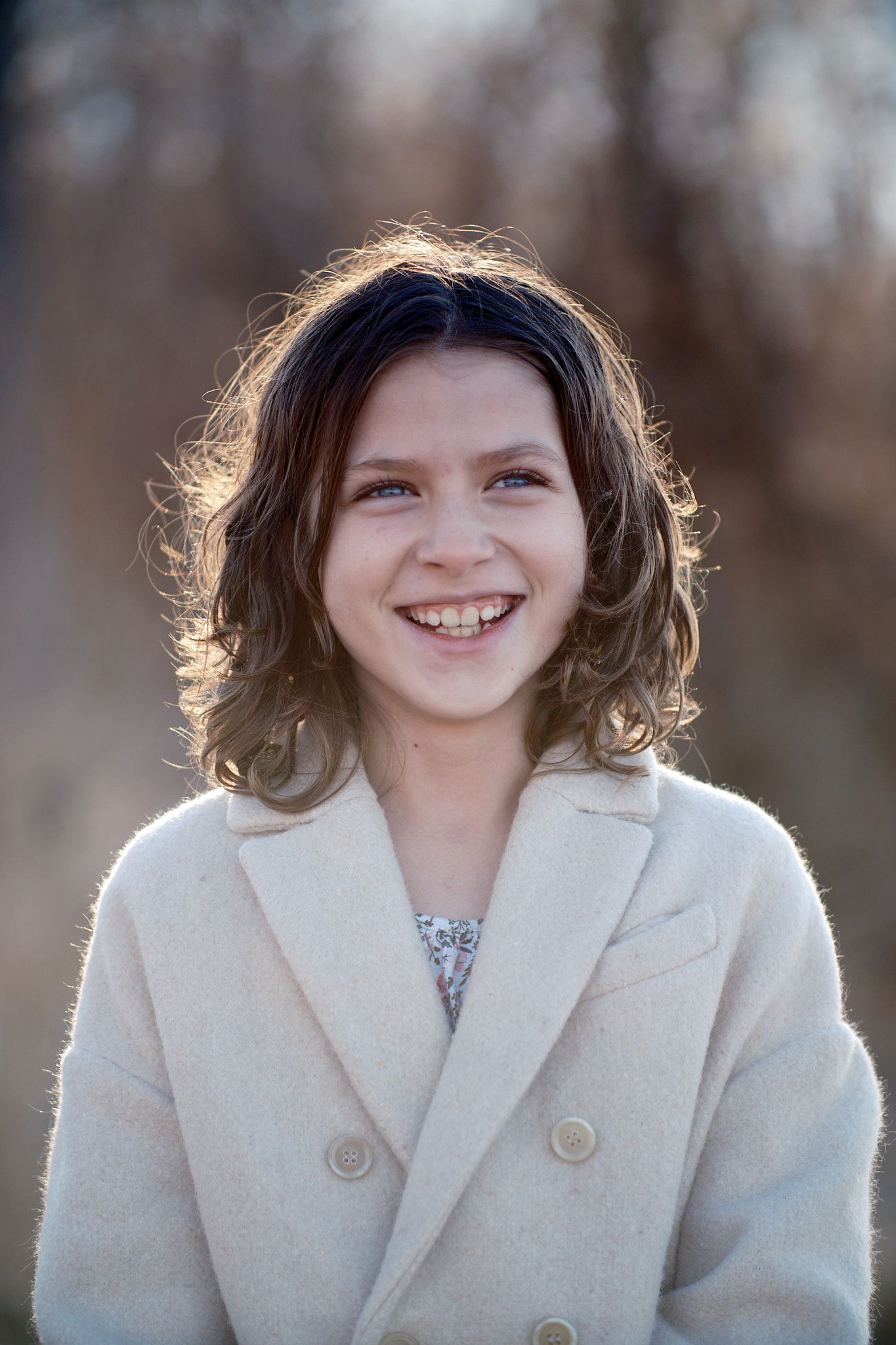 A person with curly hair smiles brightly while wearing a double-breasted cream coat, backlit by golden sunlight.