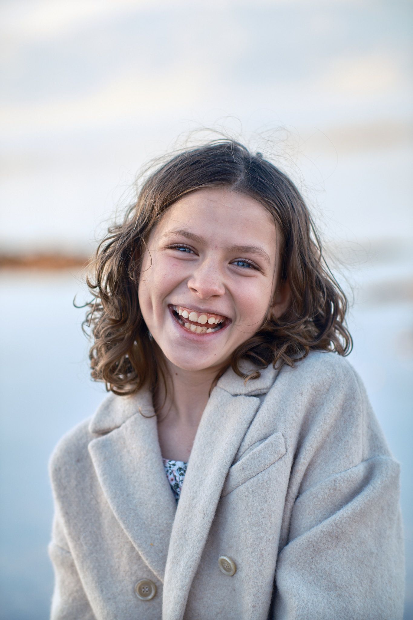 A smiling person with curly hair wearing a beige double-breasted coat against a blurred, light-colored background.