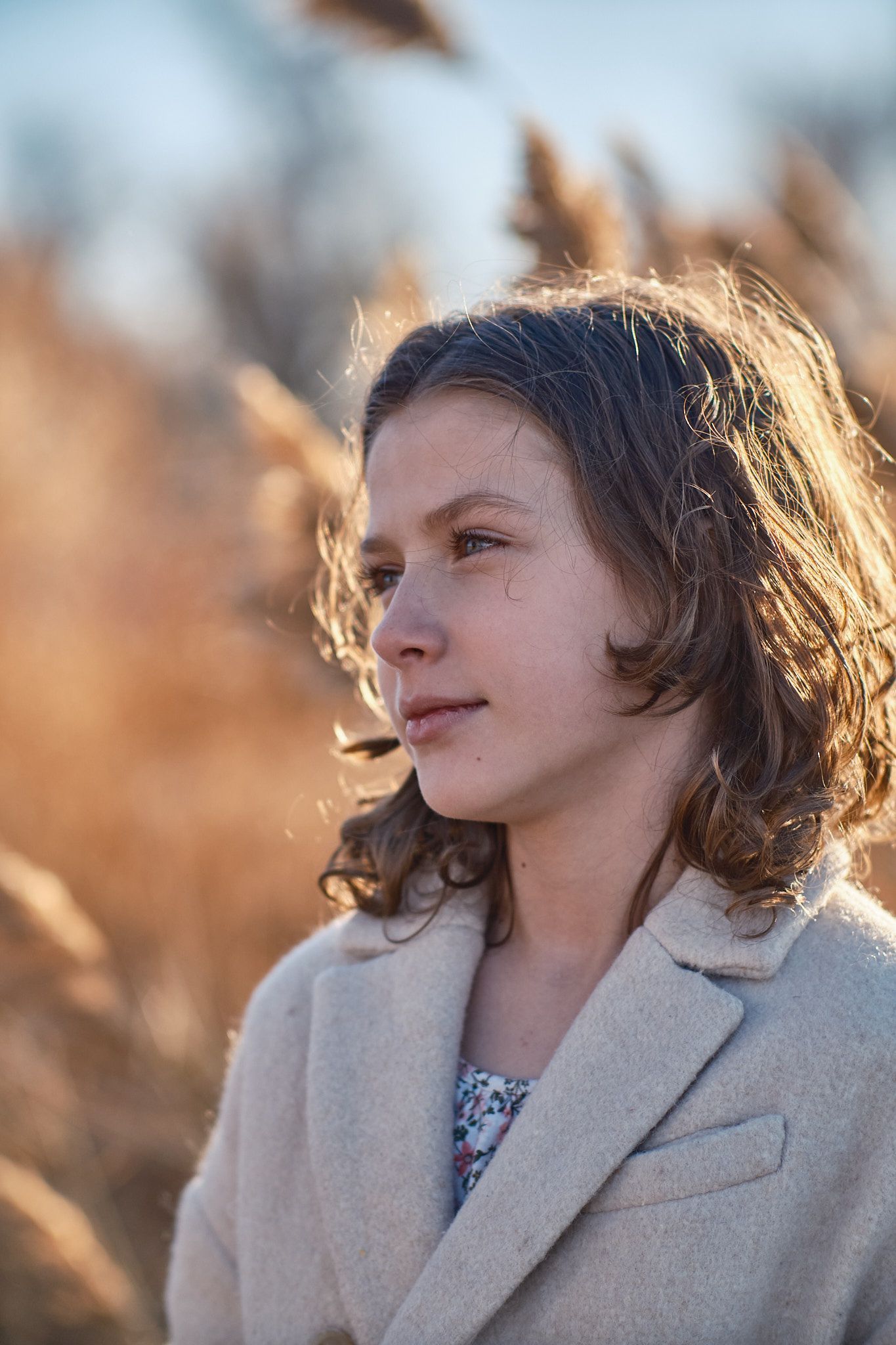 A person with wavy hair and a tan coat stands outdoors among golden tall grass, looking thoughtfully to the side.