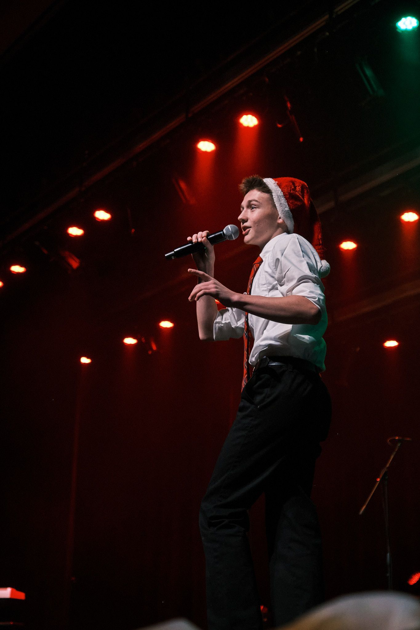 A person wearing a Santa hat and white shirt stands on a dark stage, holding a microphone and singing under red lights.