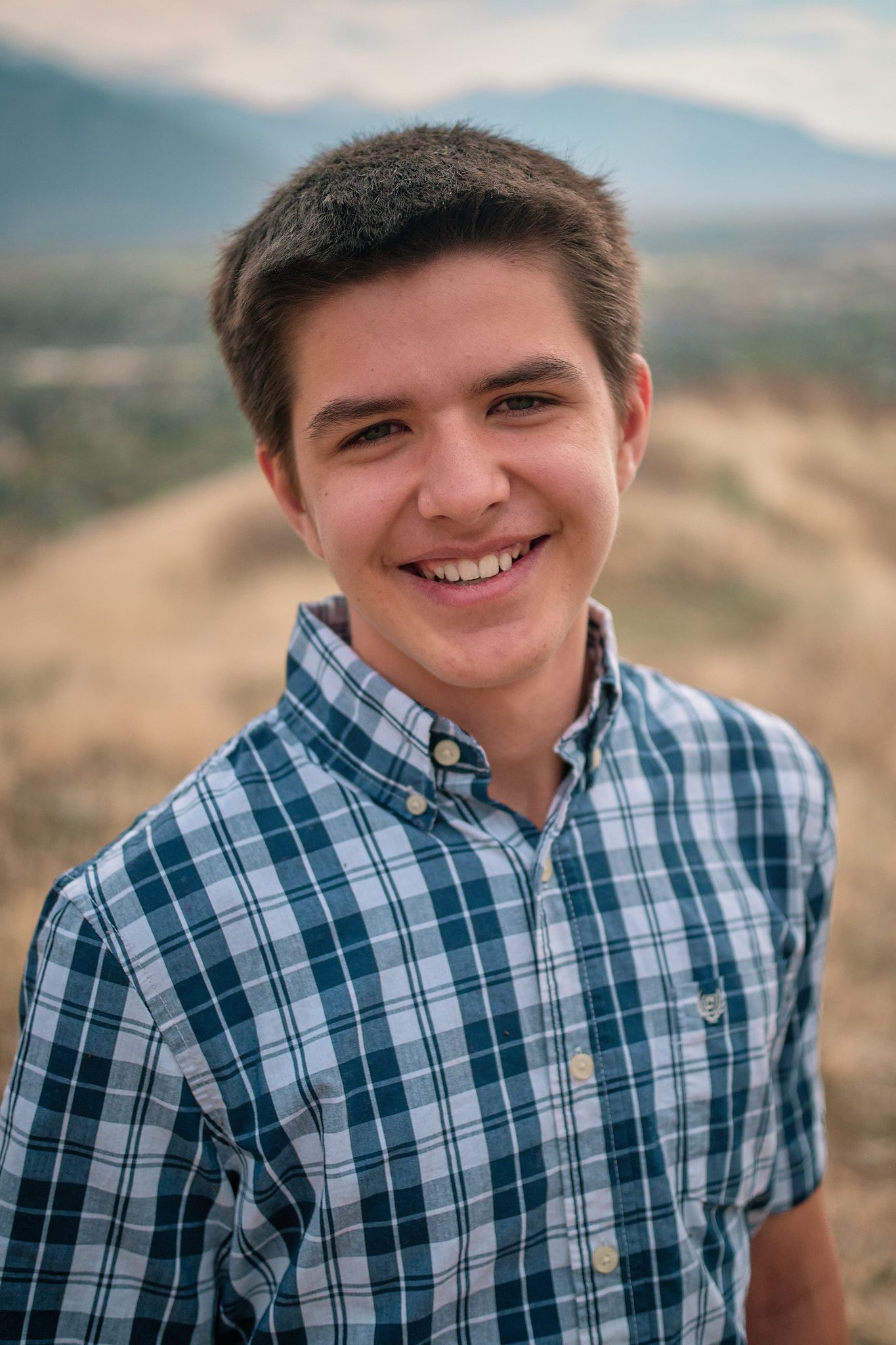 A person with short brown hair smiling, wearing a blue plaid button-down shirt outdoors against a blurred mountain backdrop.
