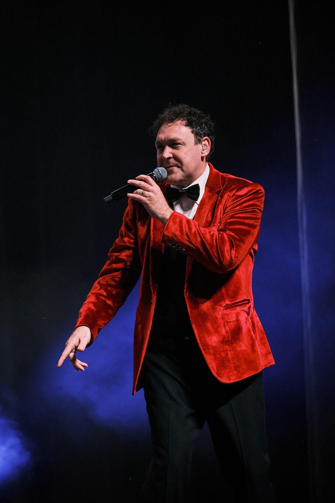 A person in a red velvet blazer and bowtie sings into a microphone on a stage with blue lighting.