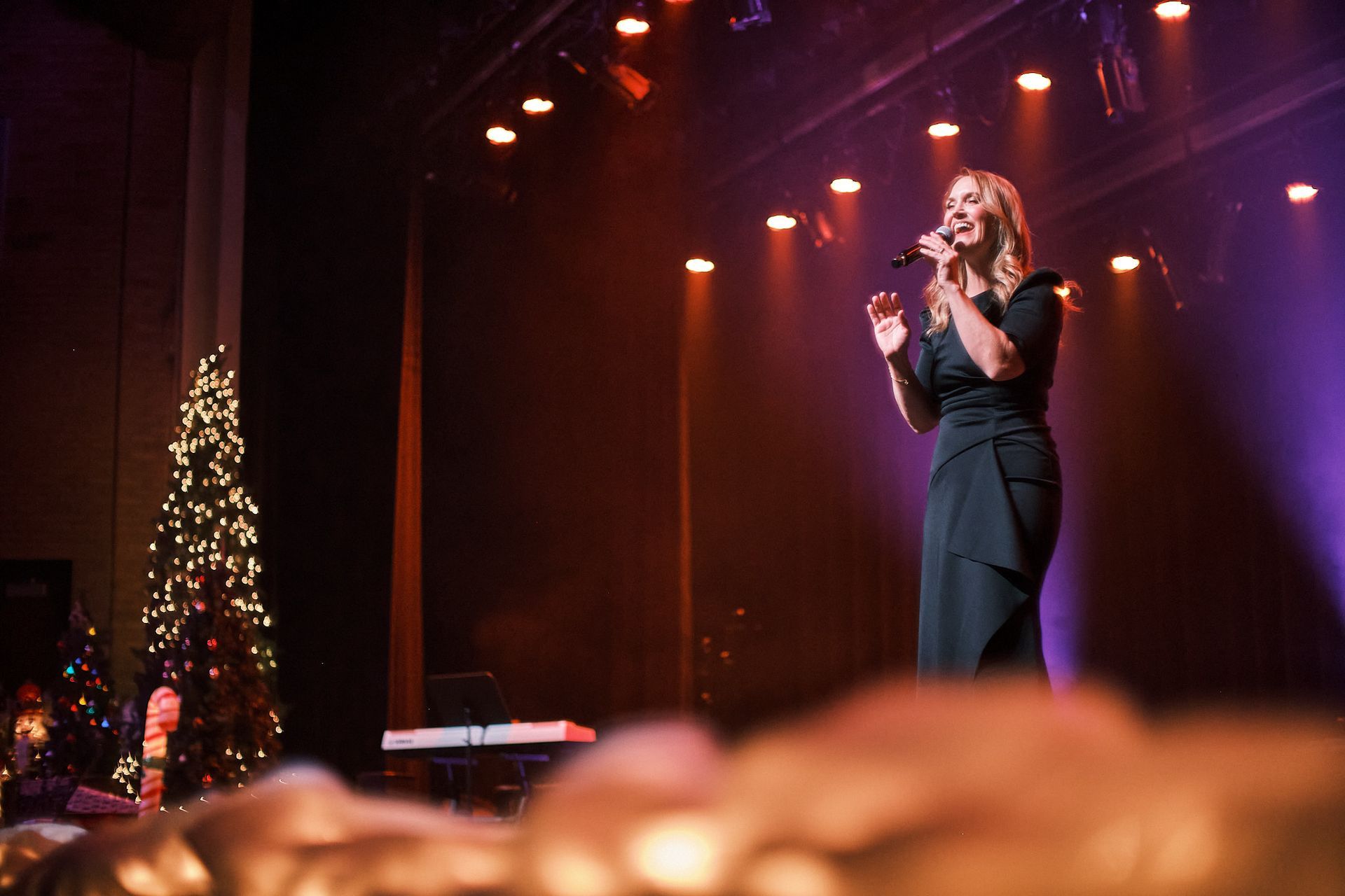 A performer in a black dress sings into a microphone on a stage beside a decorated Christmas tree.