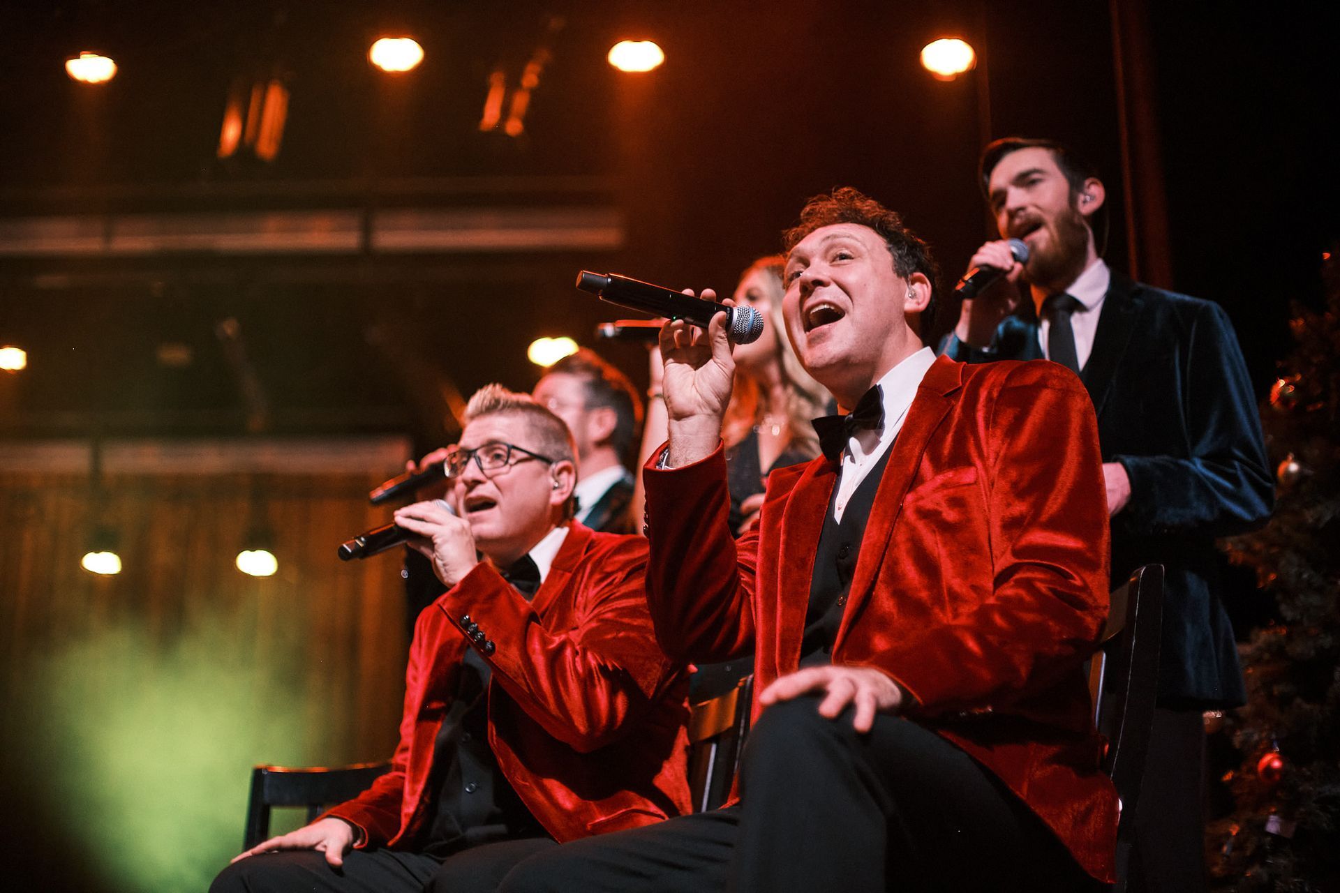 A musical group in red velvet jackets sings into microphones on a stage with warm lighting.