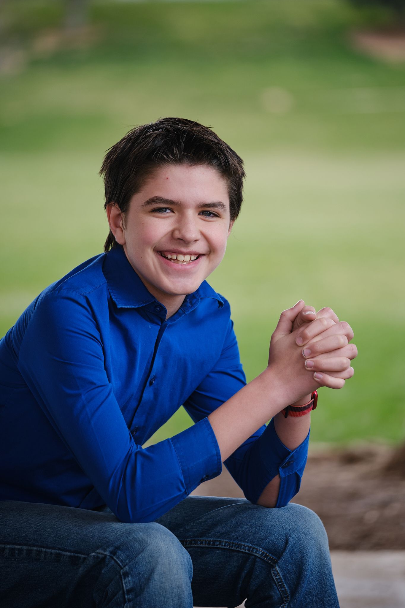 A smiling person in a blue button-down shirt and jeans sitting outdoors with hands clasped.