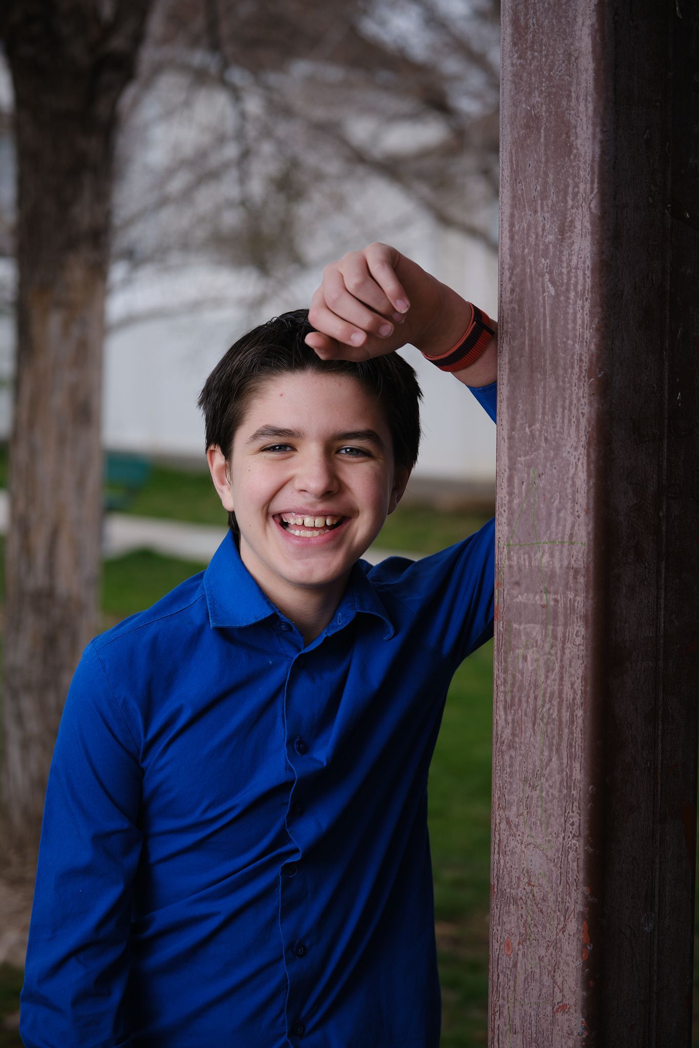 A person in a blue collared shirt smiling while leaning against a wooden post in an outdoor setting.