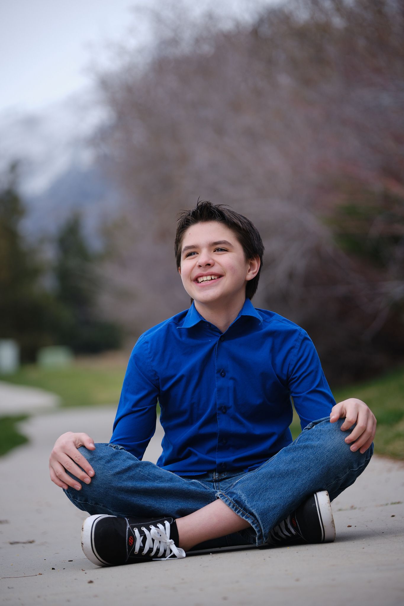 A person in a blue button-down shirt and blue jeans sits cross-legged on a paved path, looking up and smiling.