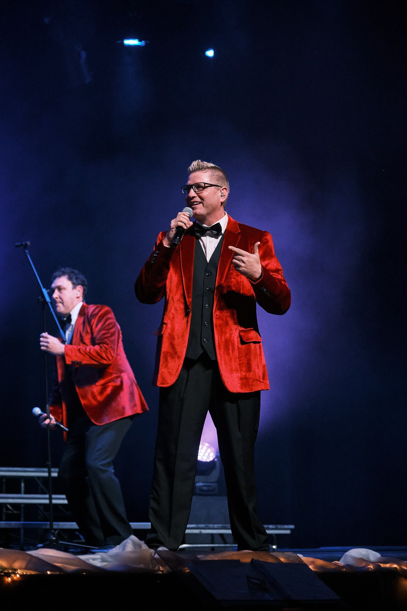 Two performers in bright red velvet jackets and black trousers sing on stage.
