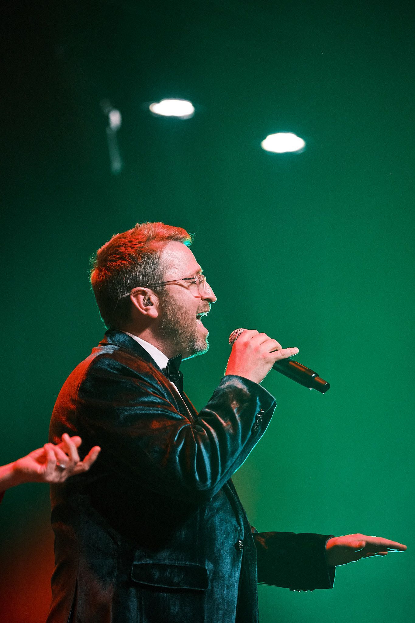 A performer with glasses sings into a microphone on stage, illuminated by green lights.