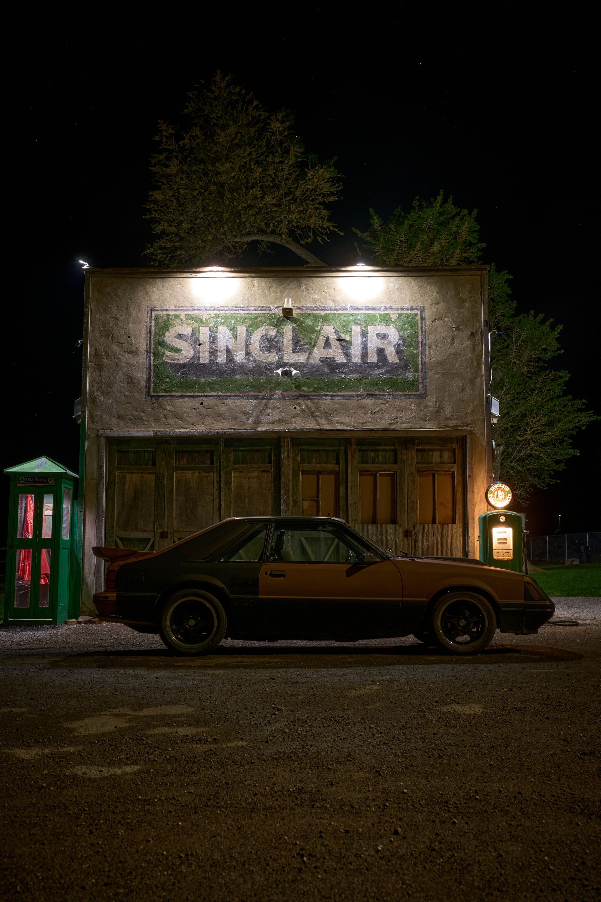 A two-tone sports car parked at night in front of an old Sinclair gas station with a telephone booth.