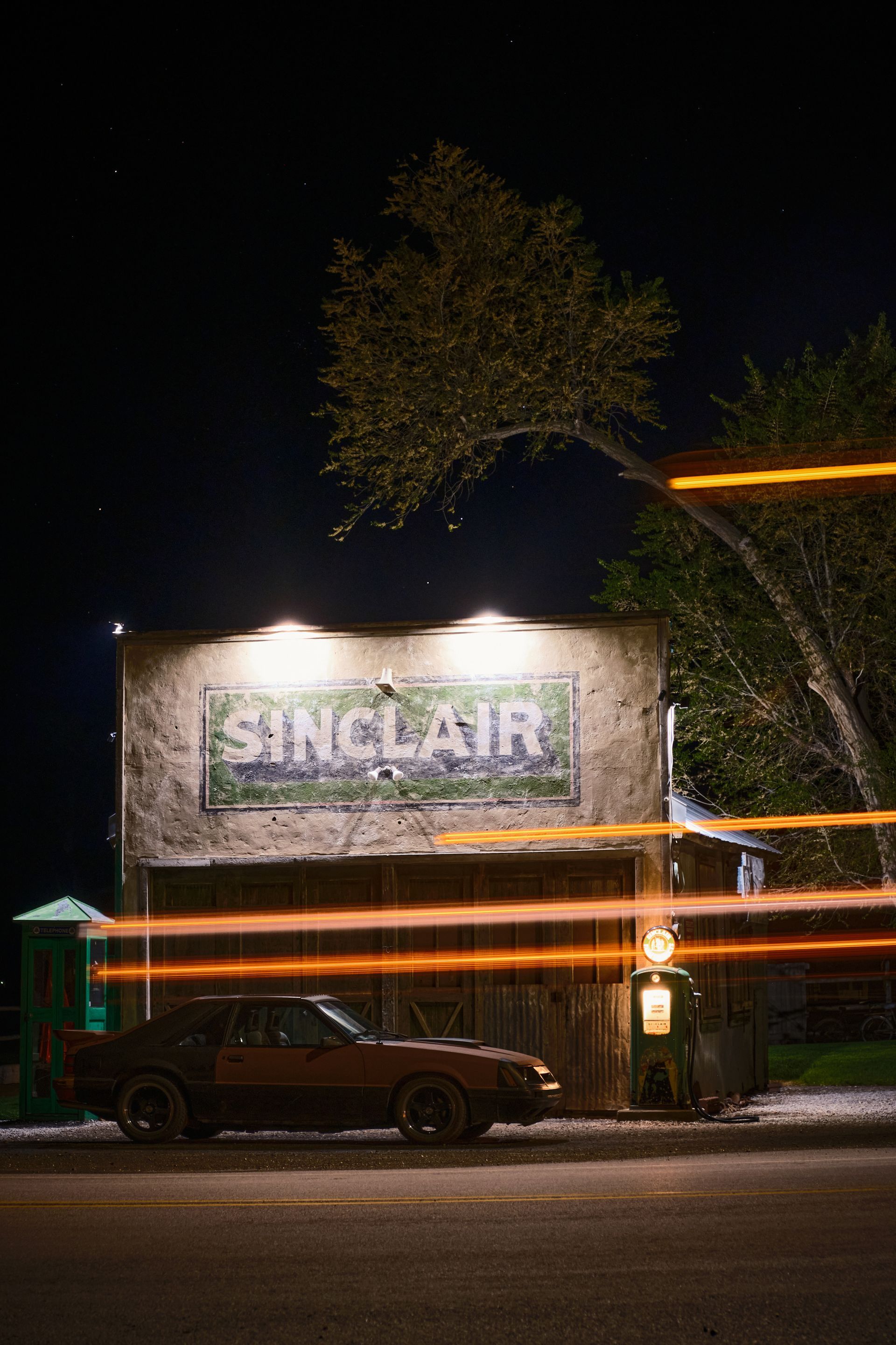 A dark, vintage Sinclair service station at night, with a car parked in front and light trails cutting across the scene.