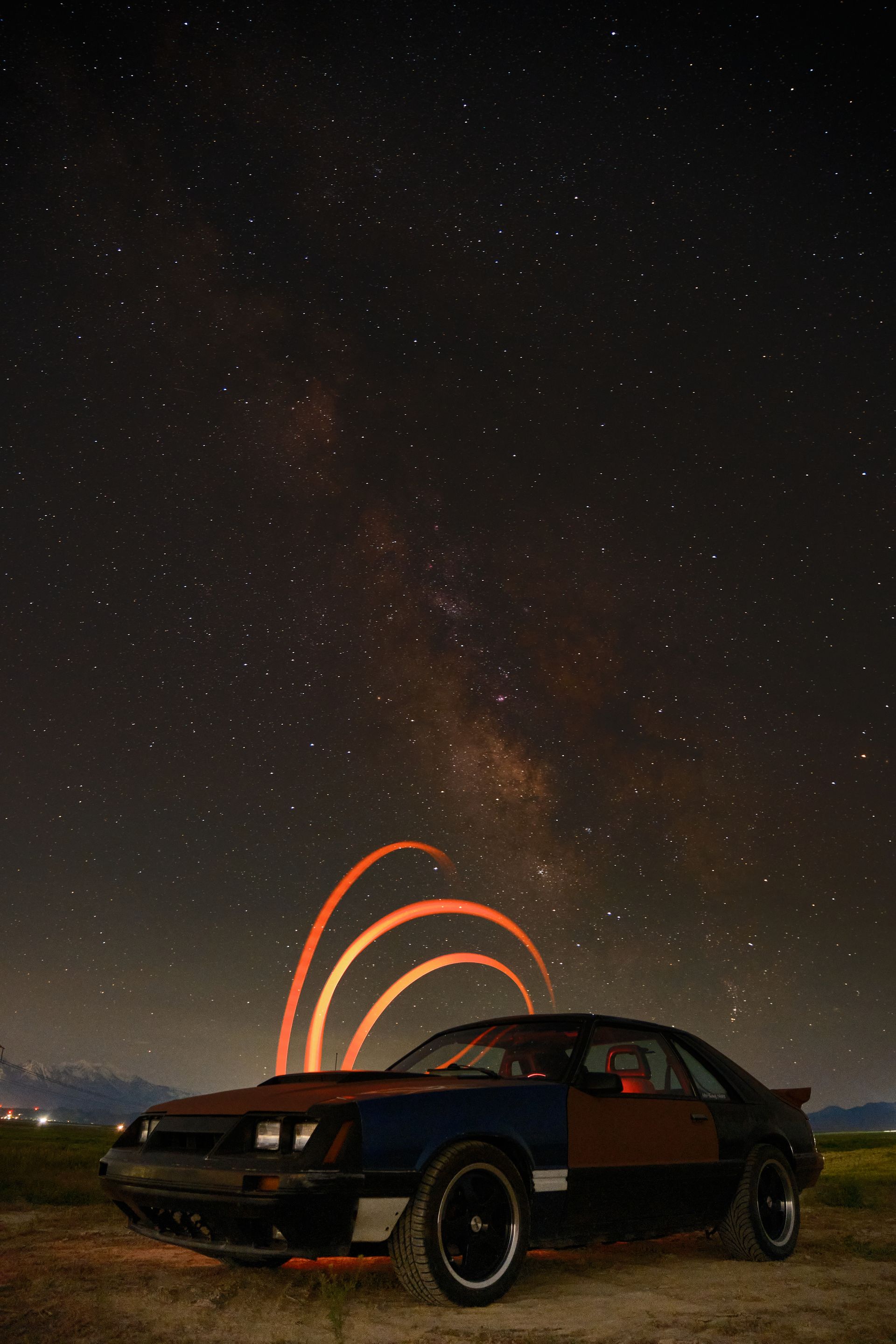 A rusty, dark-colored vintage car parked on a dirt field under a vast, starry night sky with arched light trails.
