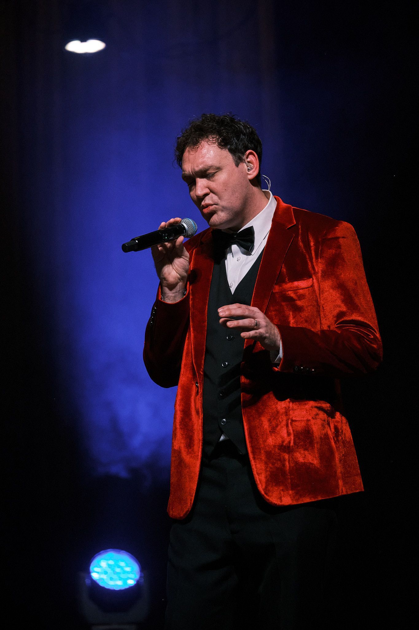 A performer in a red velvet jacket and black vest stands on a dark stage, holding a microphone and singing.
