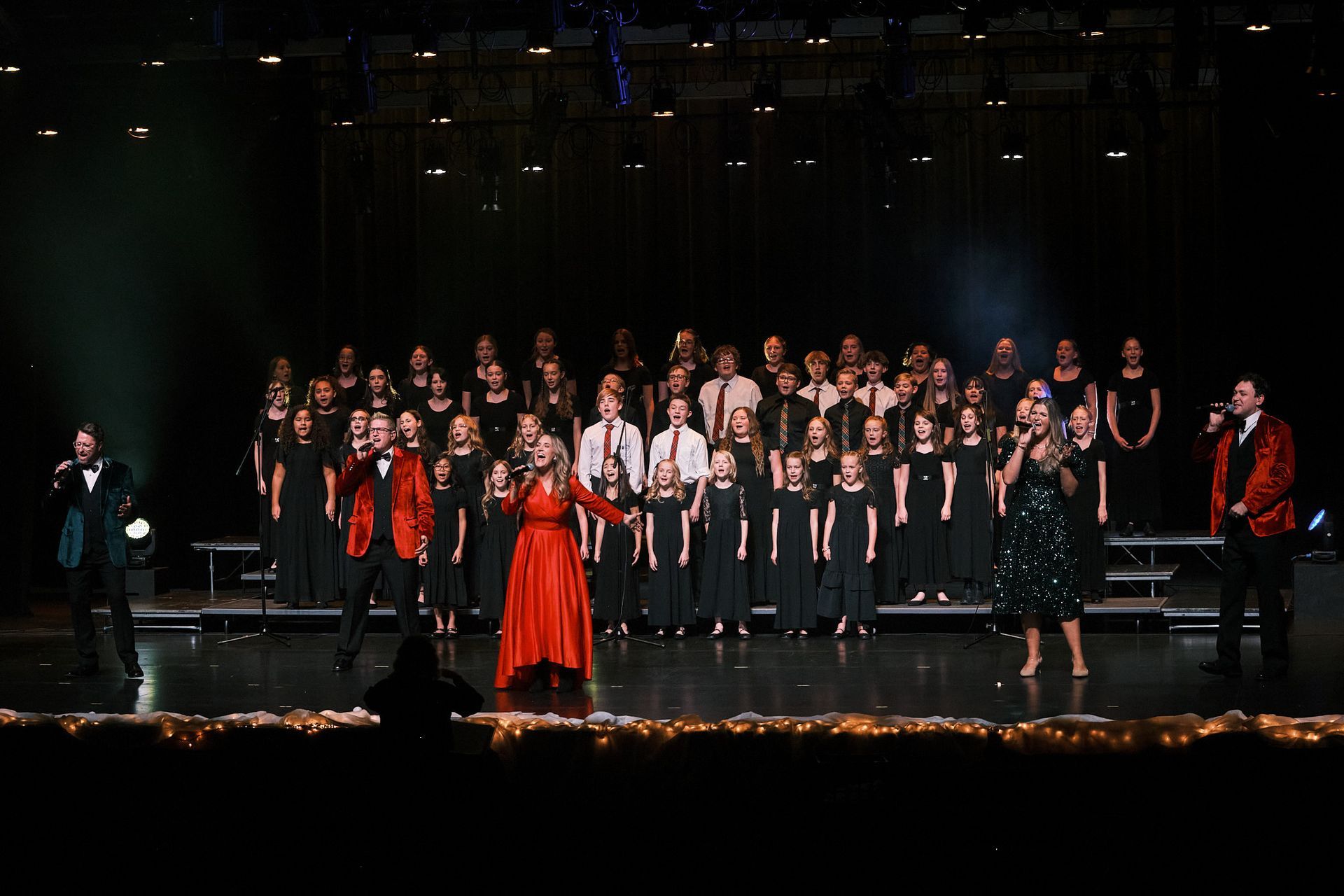 A choir performs on stage with soloists in formal wear, including red jackets and a red dress, under bright stage lights.