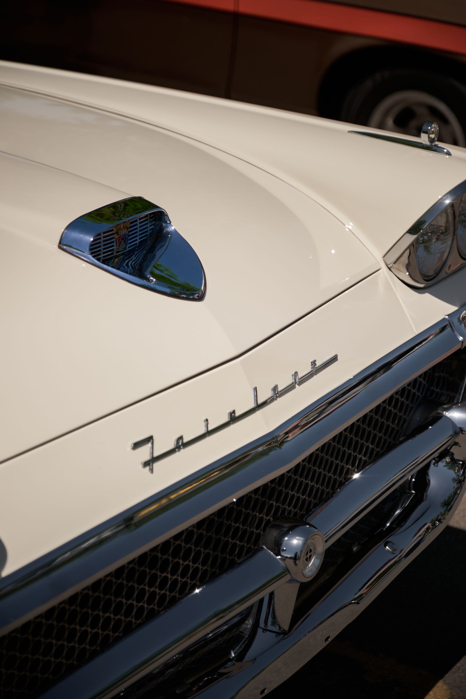 A close-up of a cream-colored vintage Ford Fairlane car hood, featuring a chrome hood ornament and grille.