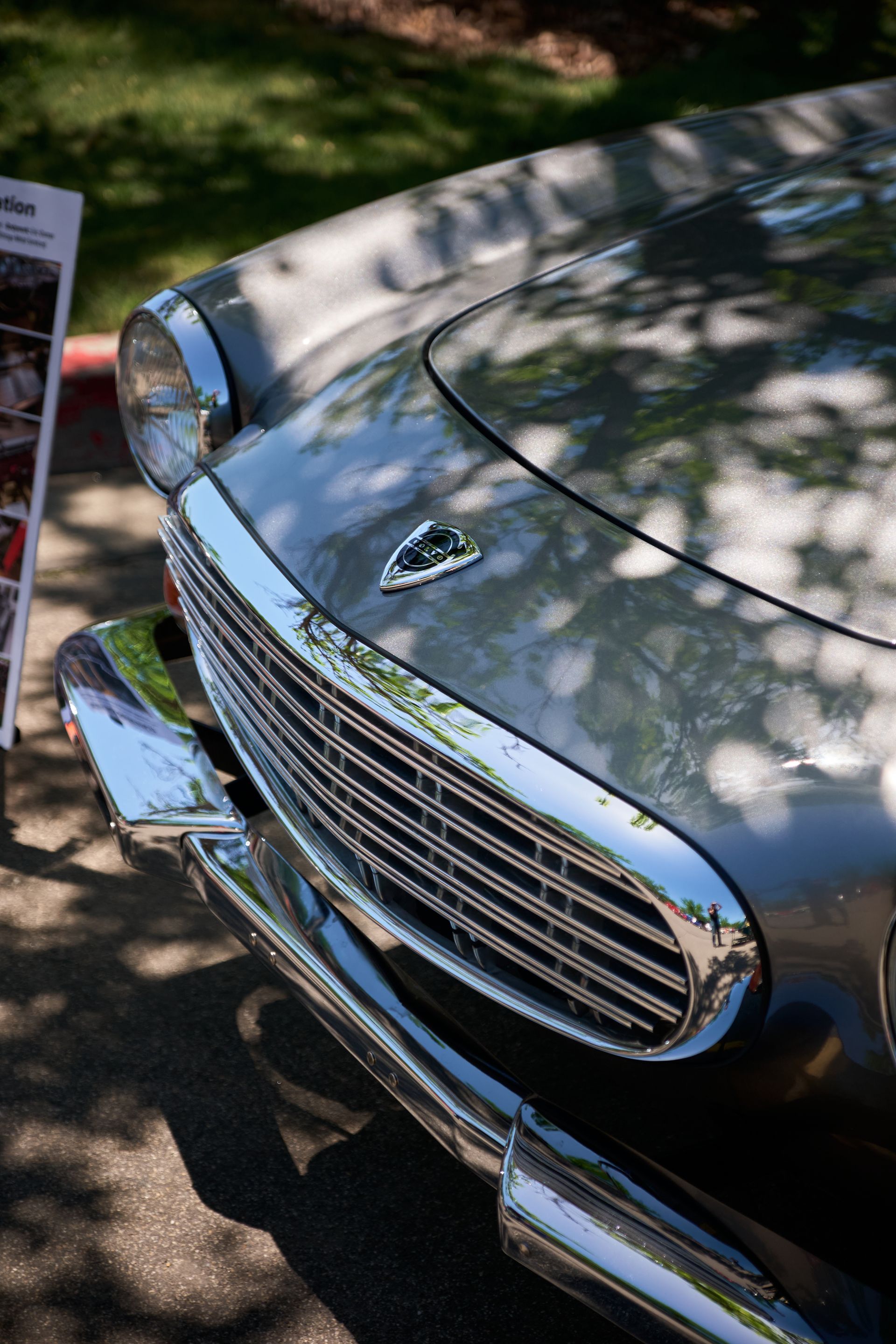 Close-up of a silver vintage car's chrome front grille, headlight, and hood emblem, dappled with sunlight and shadows.