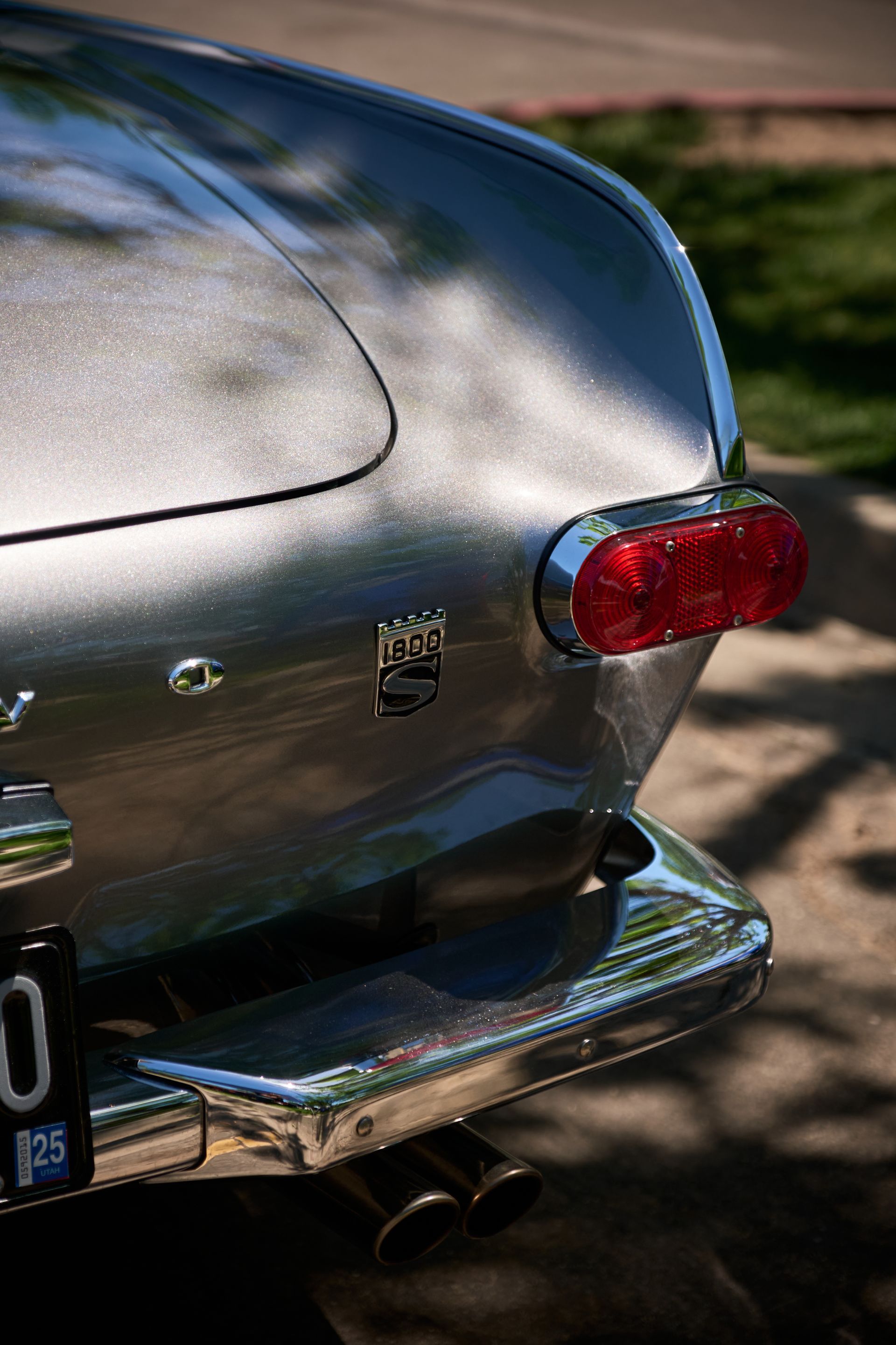 Rear view of a silver vintage Volvo P1800 with a chrome bumper, red taillight, and dual exhaust pipes on an outdoor road.