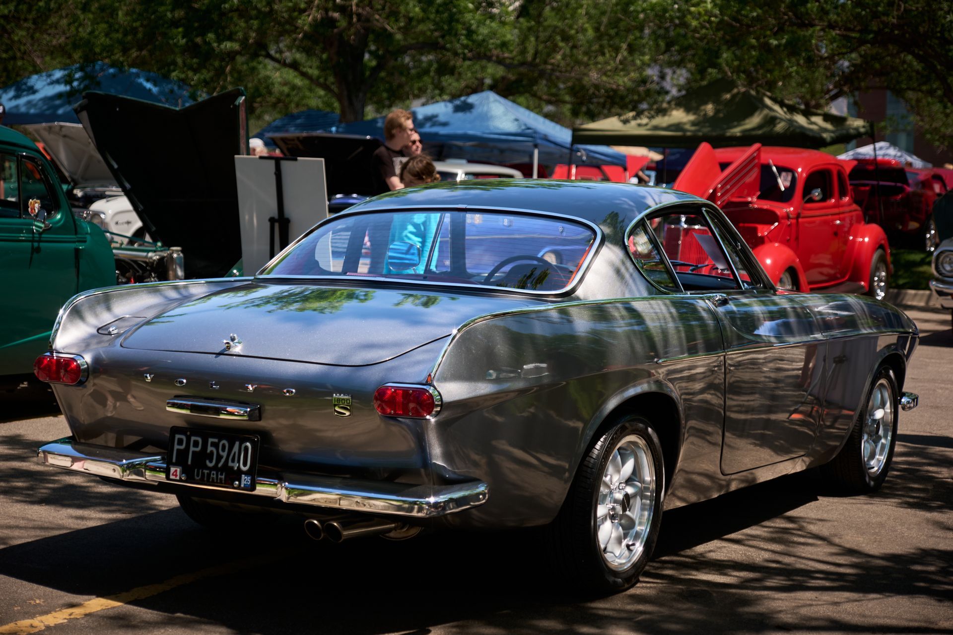 A silver Volvo P1800 coupe parked at an outdoor car show, viewed from the rear three-quarter angle.