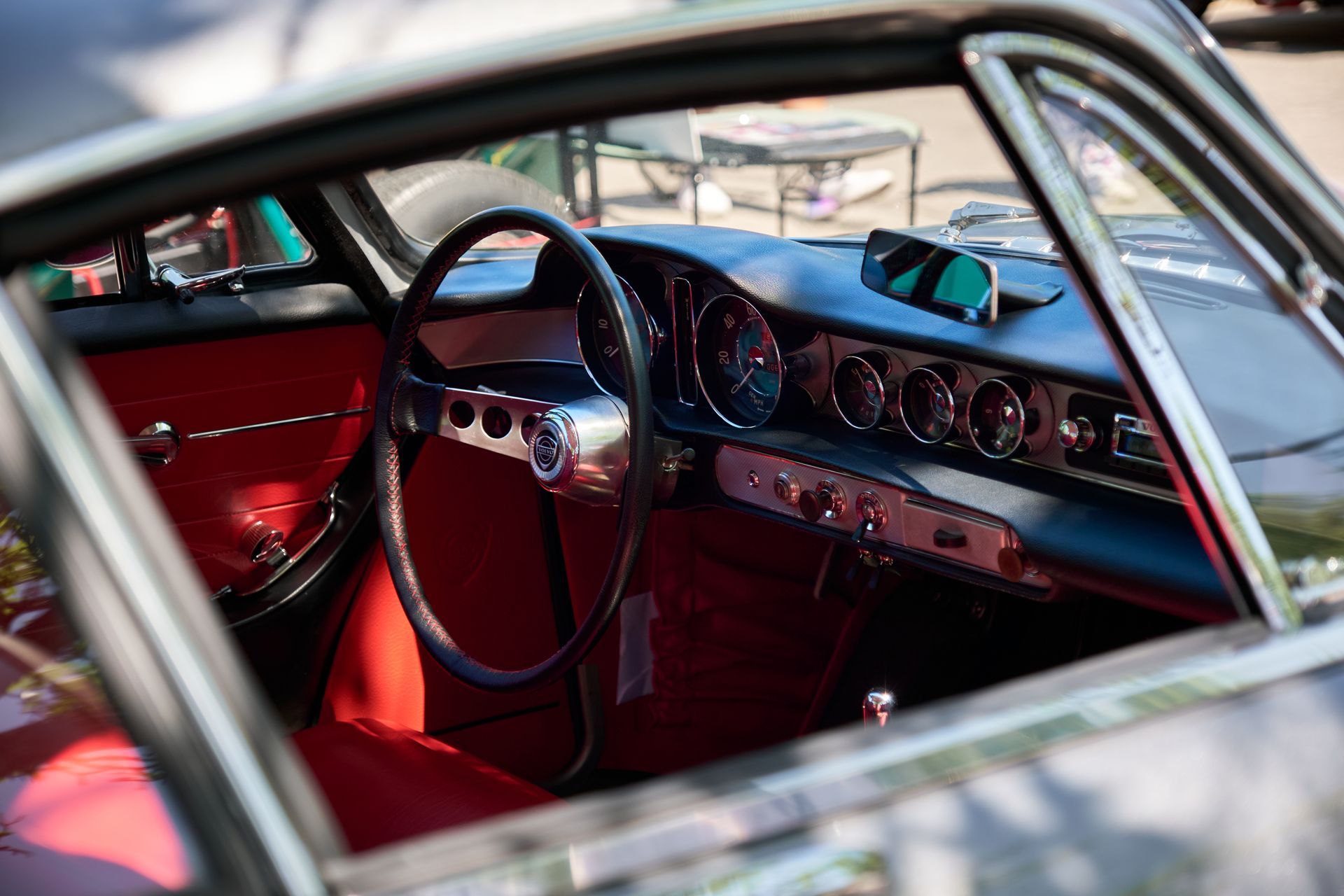 A classic car interior features red upholstery, a spoked steering wheel, and a dashboard with multiple circular gauges.