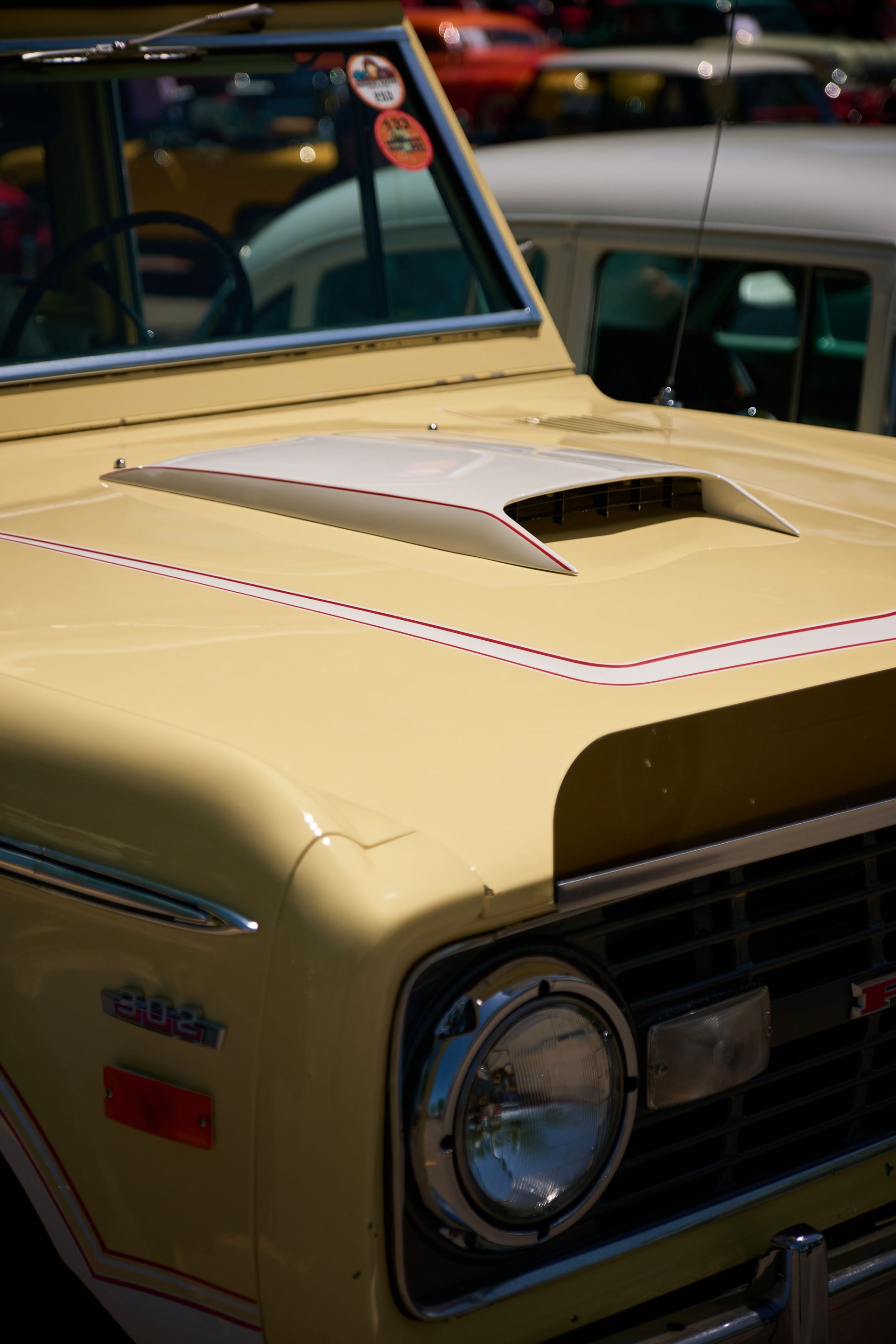 A light yellow vintage Ford Bronco, showing the hood, headlight, and a white scoop, parked at an outdoor car show.