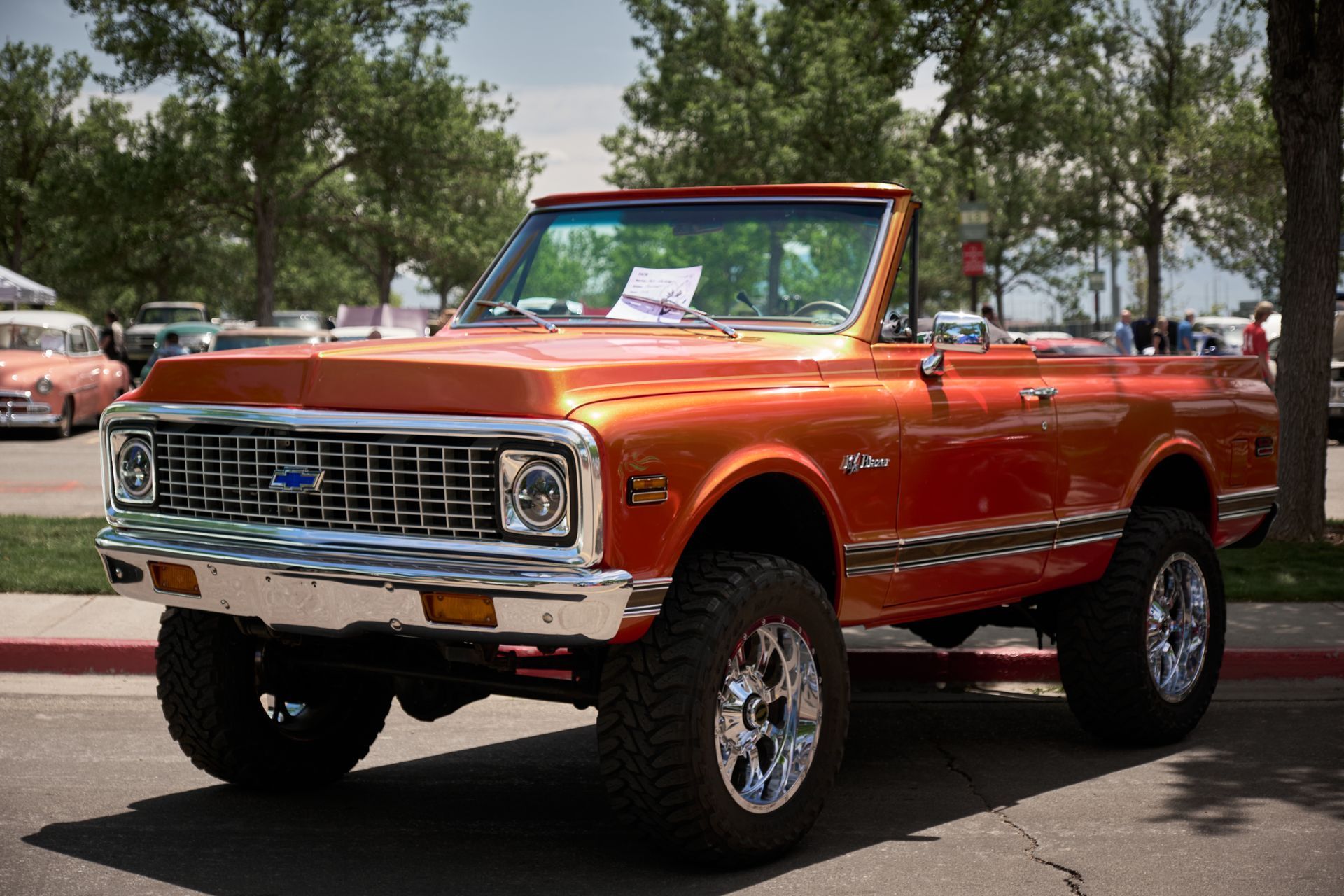 An orange vintage Chevrolet Blazer convertible with a lifted suspension and chrome wheels, parked at an outdoor car show.