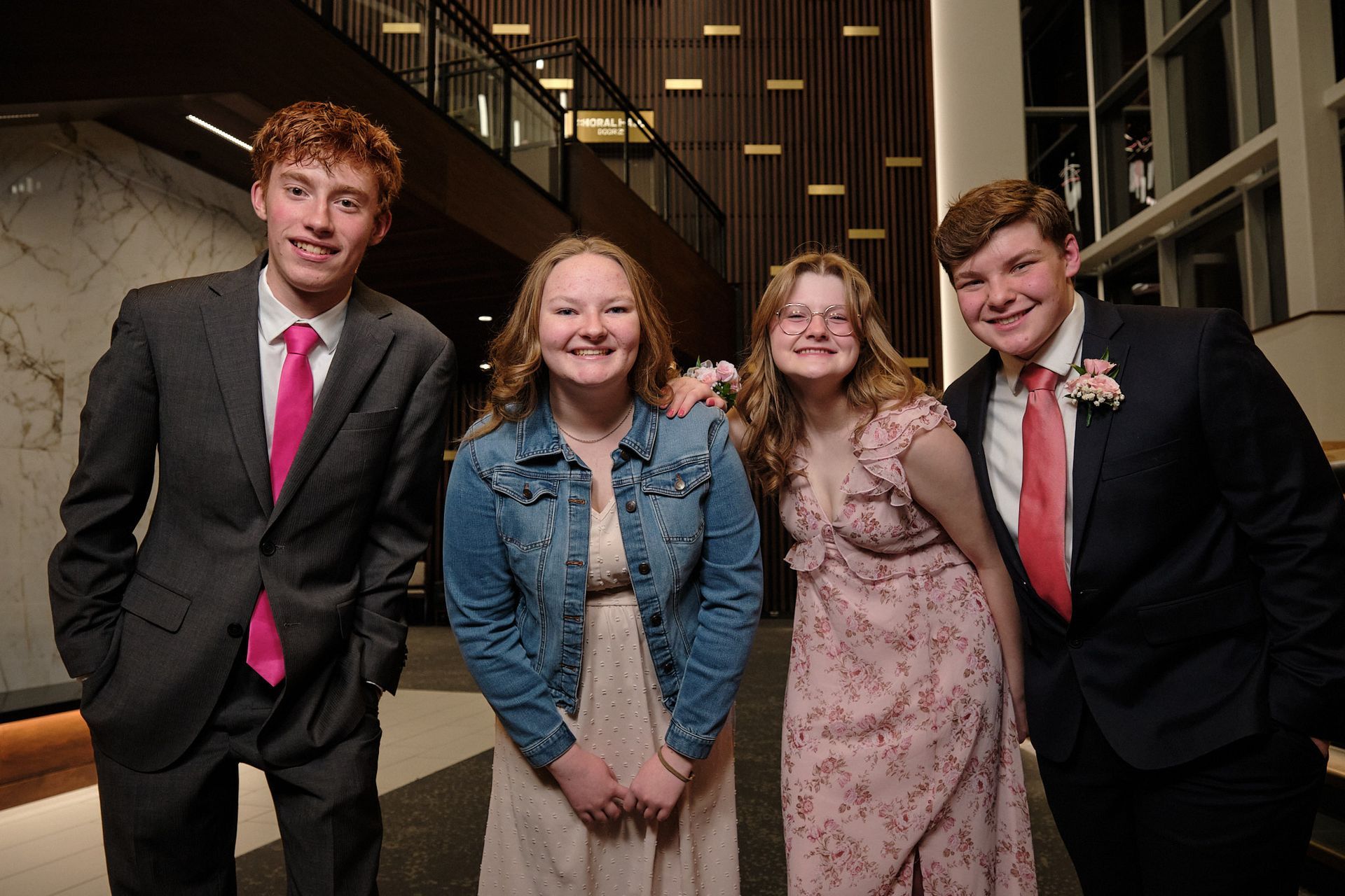 Two people in formal suits and two in dresses pose smiling together in a brightly lit indoor space with wood paneling.