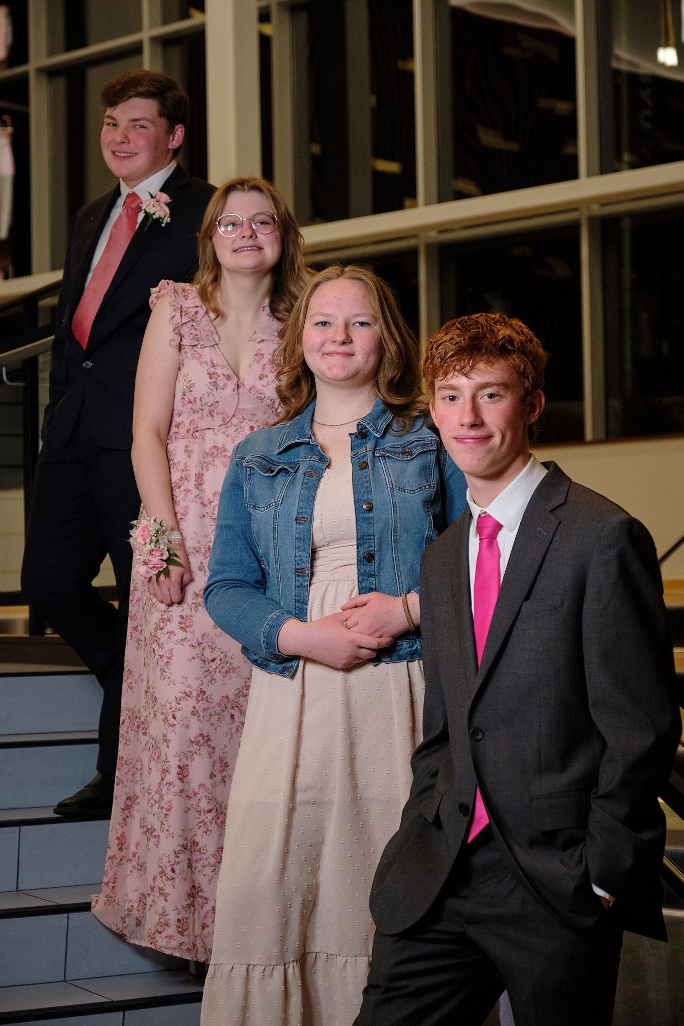 Two people in formal wear and two people in semi-formal dresses stand on stairs inside a building with glass windows.