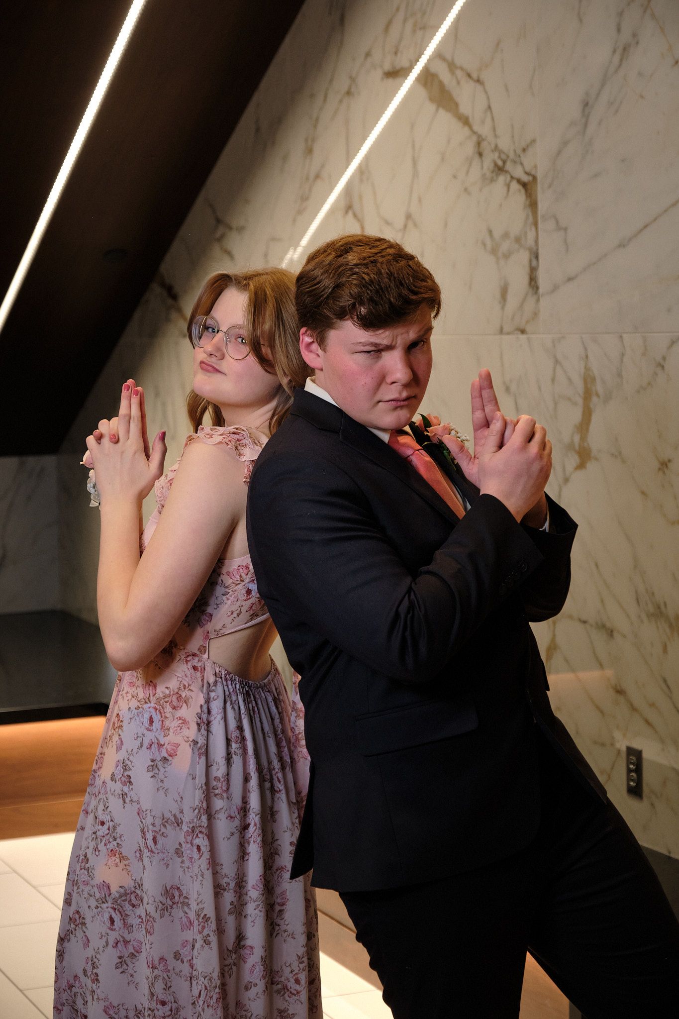A person in a floral gown and a person in a suit stand back-to-back, posing with hands shaped like pistols.