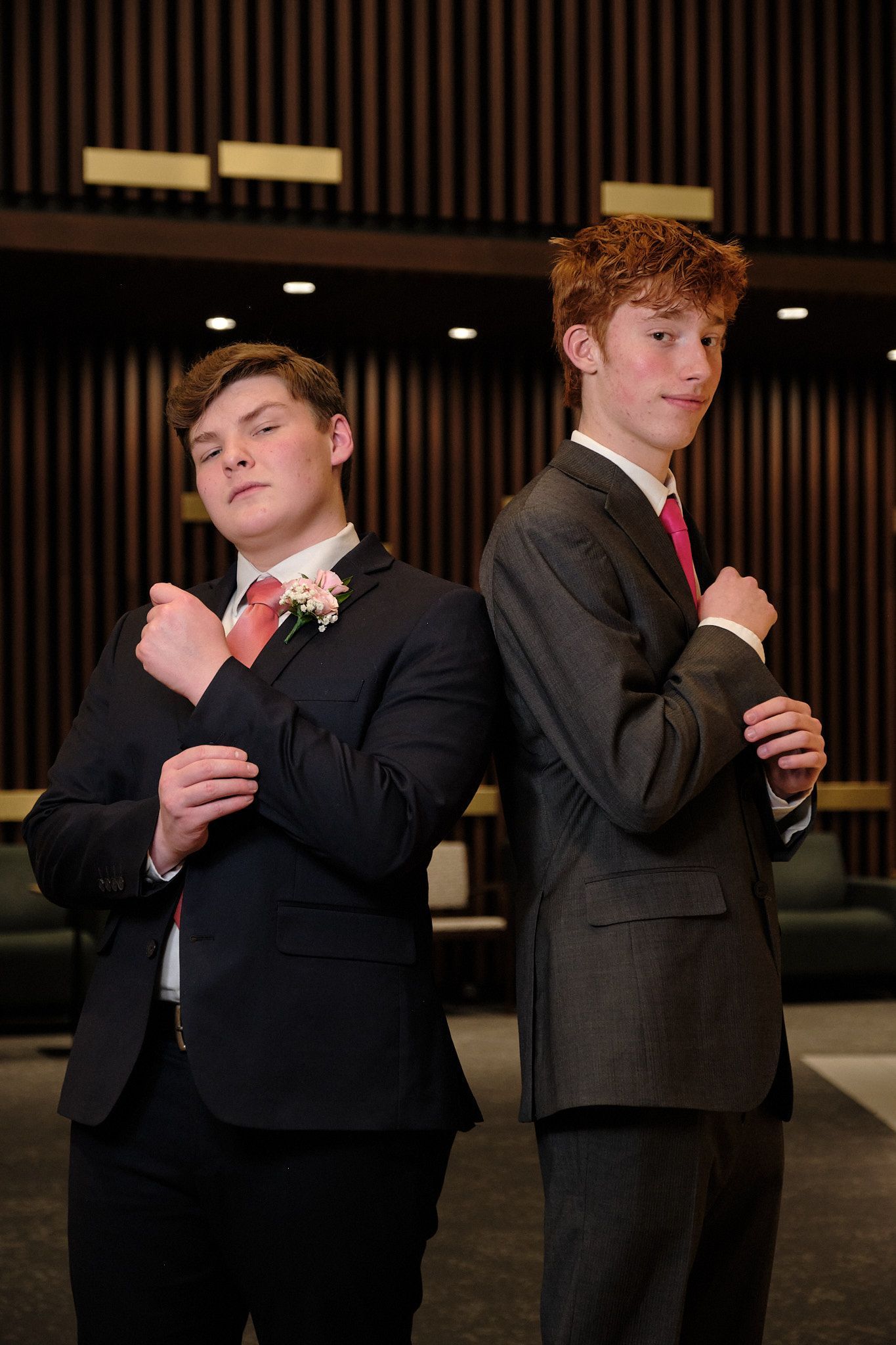 Two young men in suits and pink ties stand back-to-back, adjusting their cuffs in a modern, wood-paneled room.