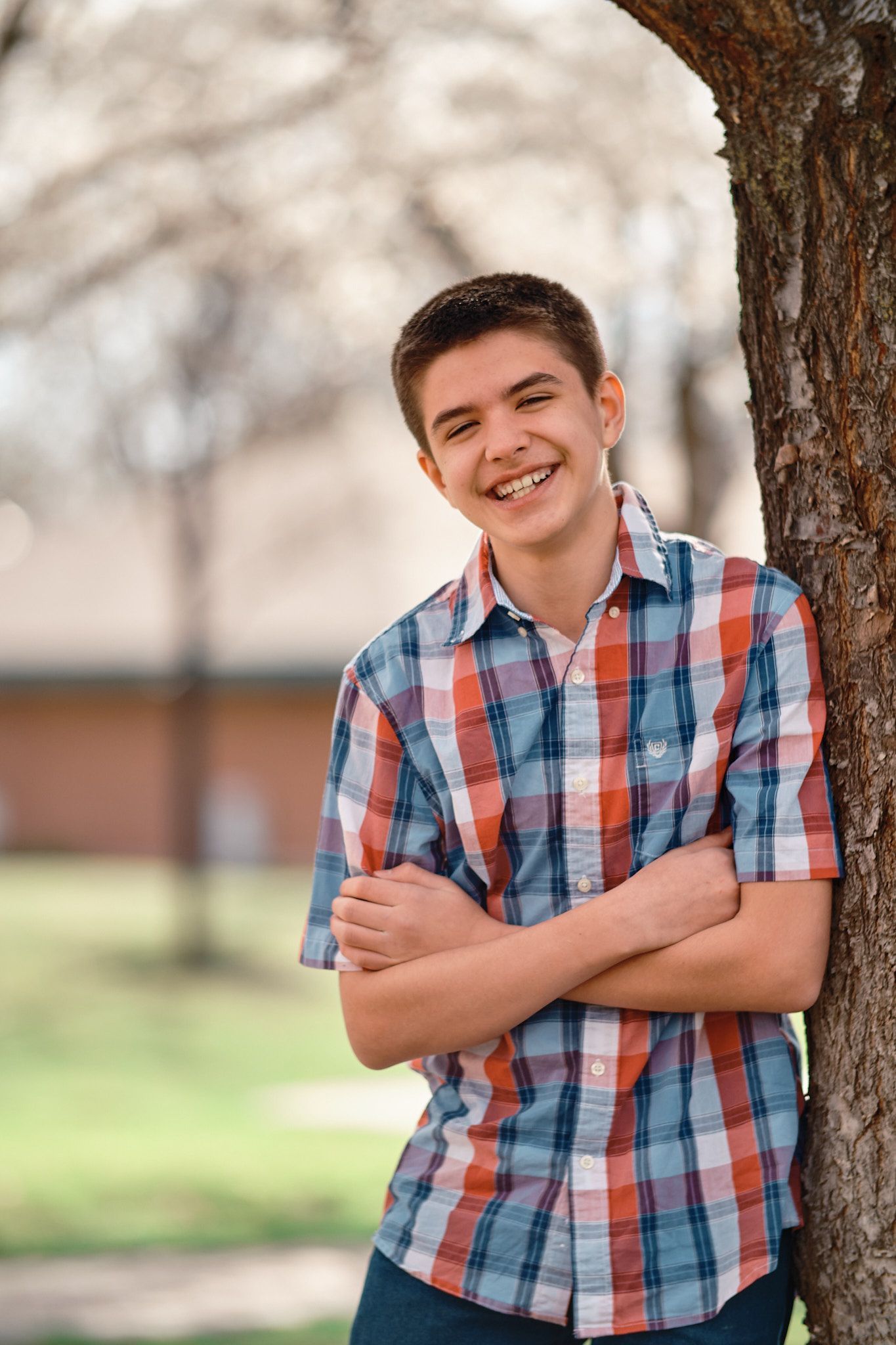 A person wearing a plaid shirt smiles with arms crossed, leaning against a tree in a park setting.