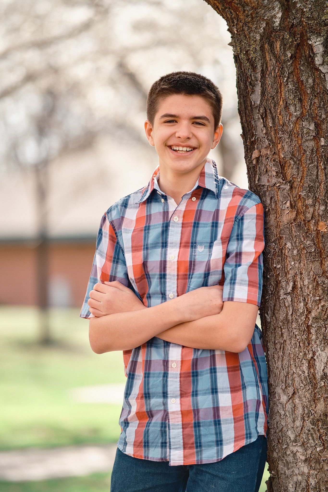 A smiling person with arms crossed, leaning against a tree in a park with a soft-focus background.