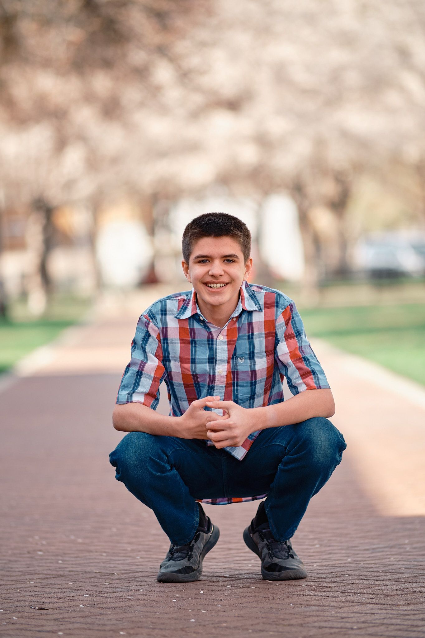 A smiling person wearing a plaid shirt and blue jeans, squatting on a paved path lined with trees.