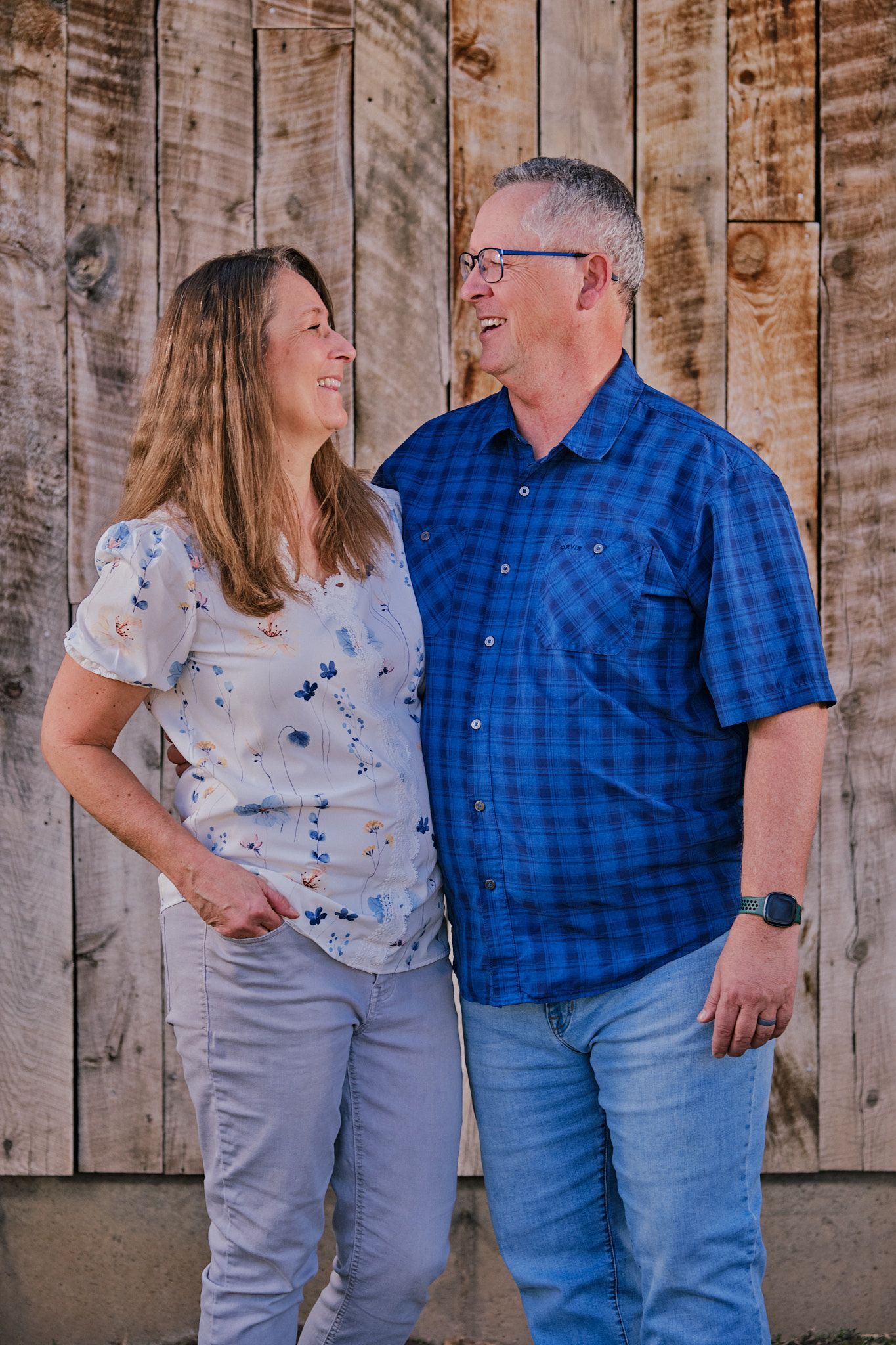 A smiling couple stands side-by-side against a wooden background, looking at each other.