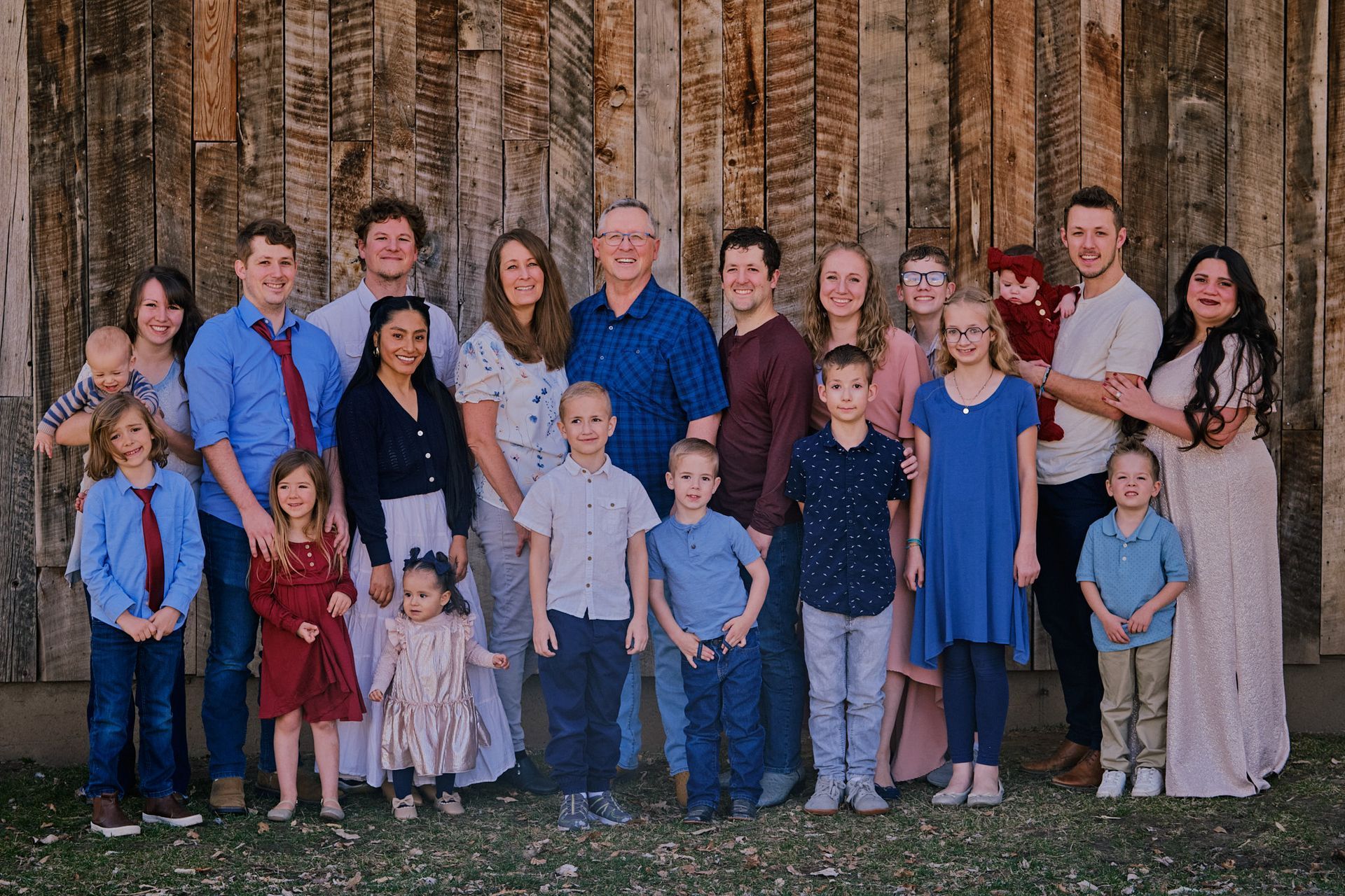 A large, multi-generational family poses outdoors in front of a weathered wood wall.
