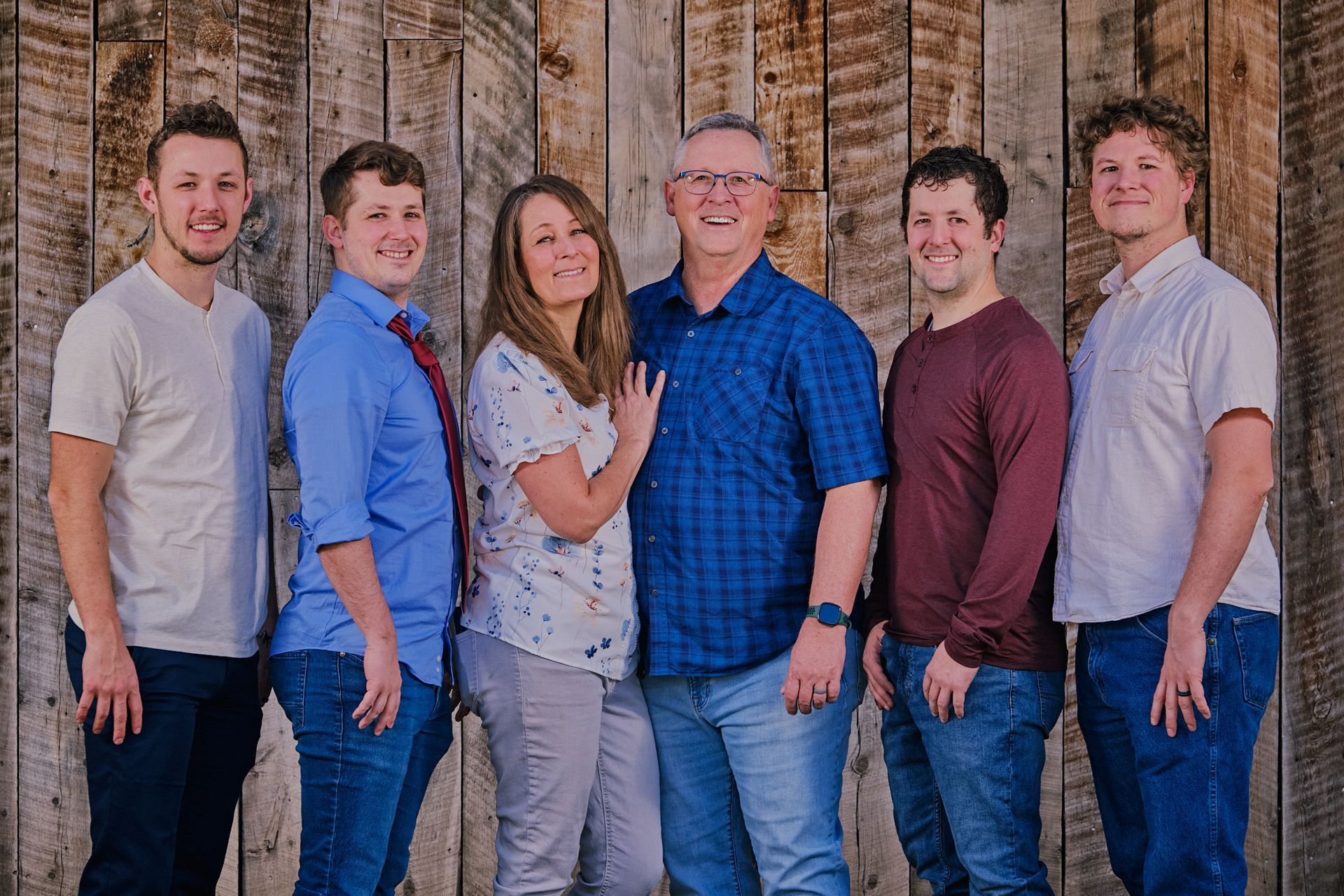 A family of six stands in a row smiling in front of a rustic wooden plank wall.