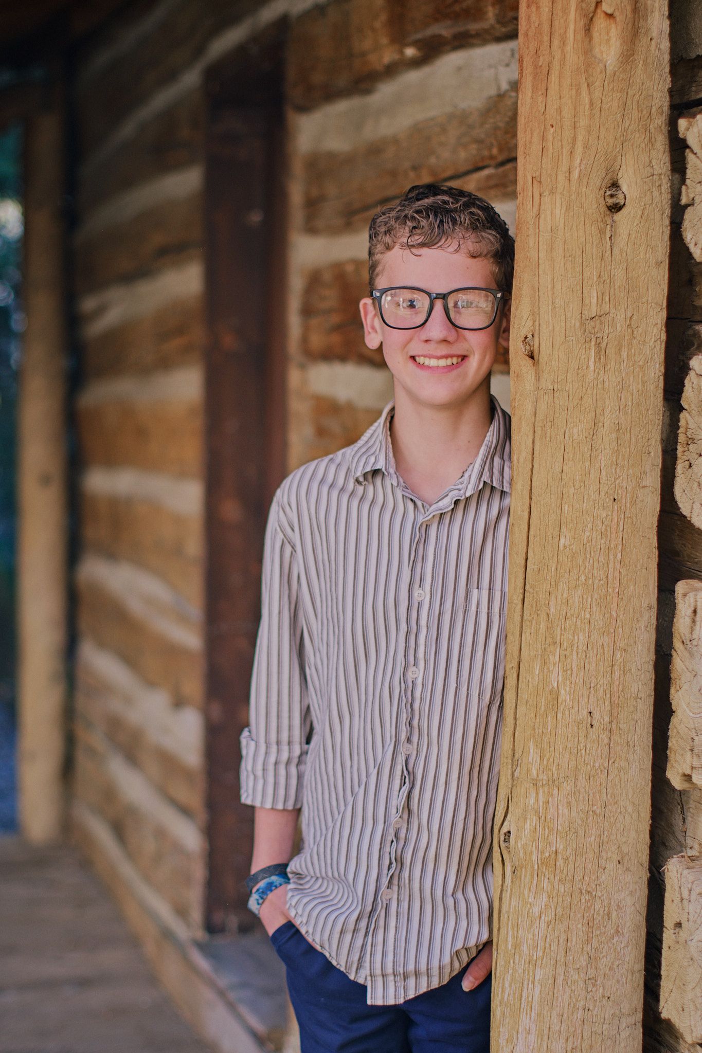 A smiling person wearing glasses and a patterned button-down shirt stands against a rustic log wall.