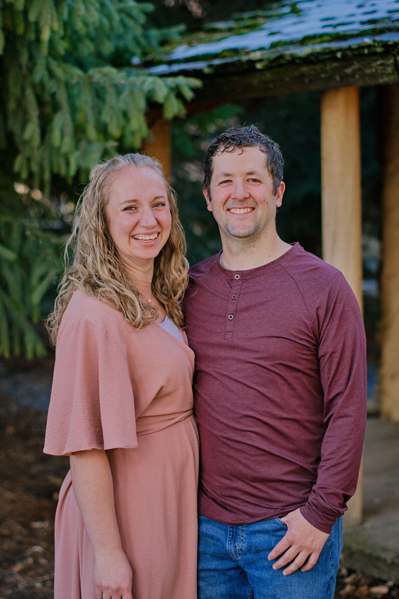 A smiling couple stands side-by-side outdoors in front of a tree and a wooden structure.