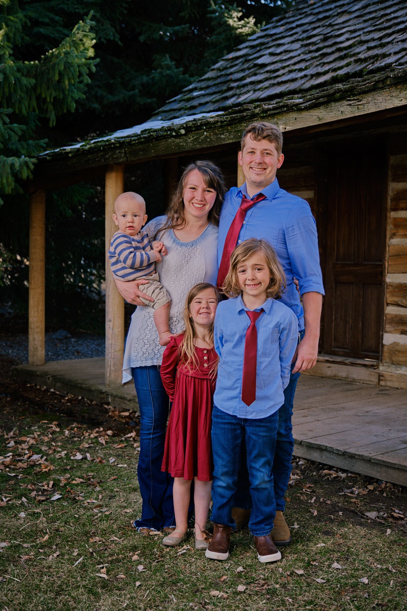 A family stands together in front of a rustic log cabin, dressed in blue shirts and jeans, with one in a red dress.