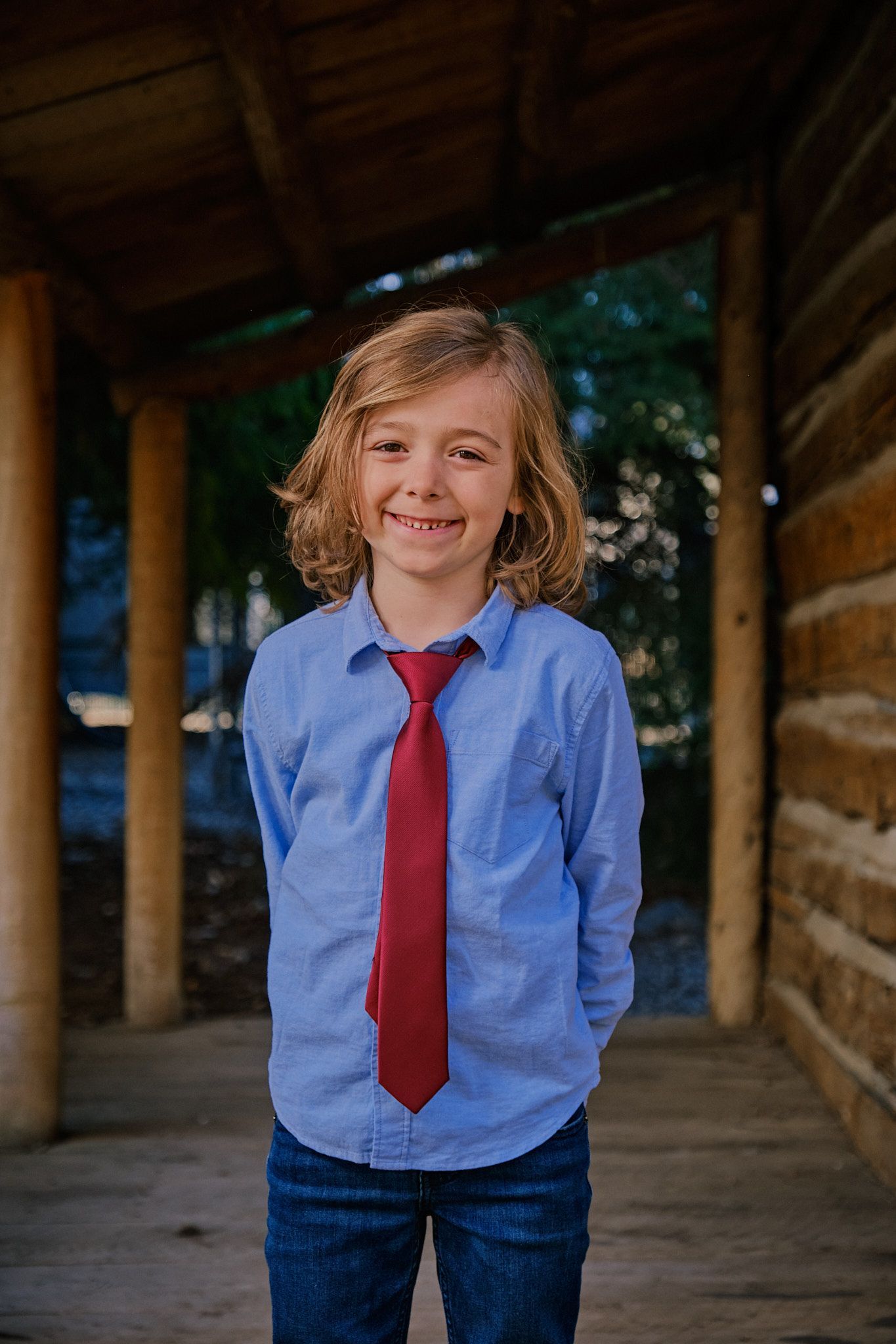 A person with shoulder-length wavy hair, wearing a blue button-down shirt and red tie, smiles on a wooden porch.