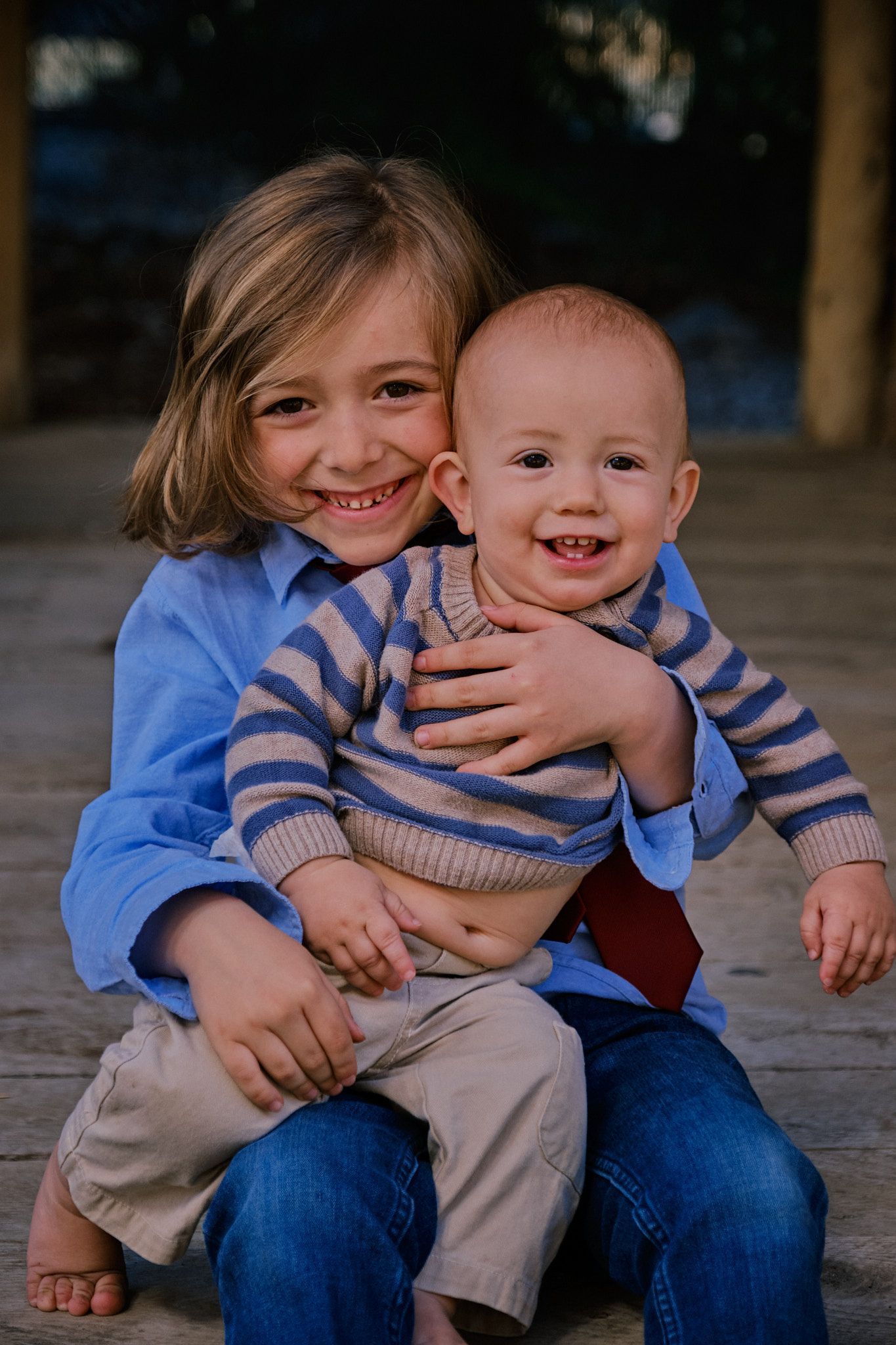 An older child in a blue shirt holds a smiling baby wearing a blue and grey striped sweater on their lap.