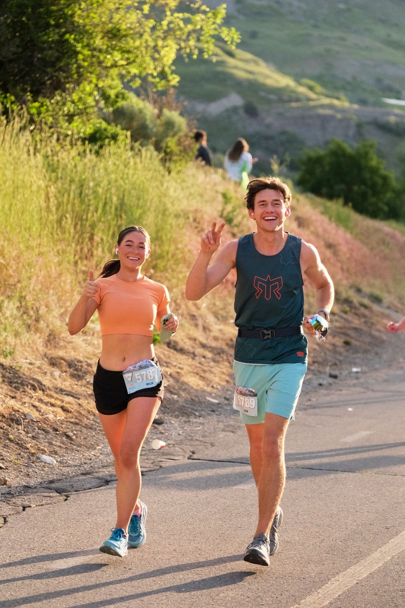 Two runners smile and make hand gestures while jogging on a sunny, paved path beside a grassy hillside.